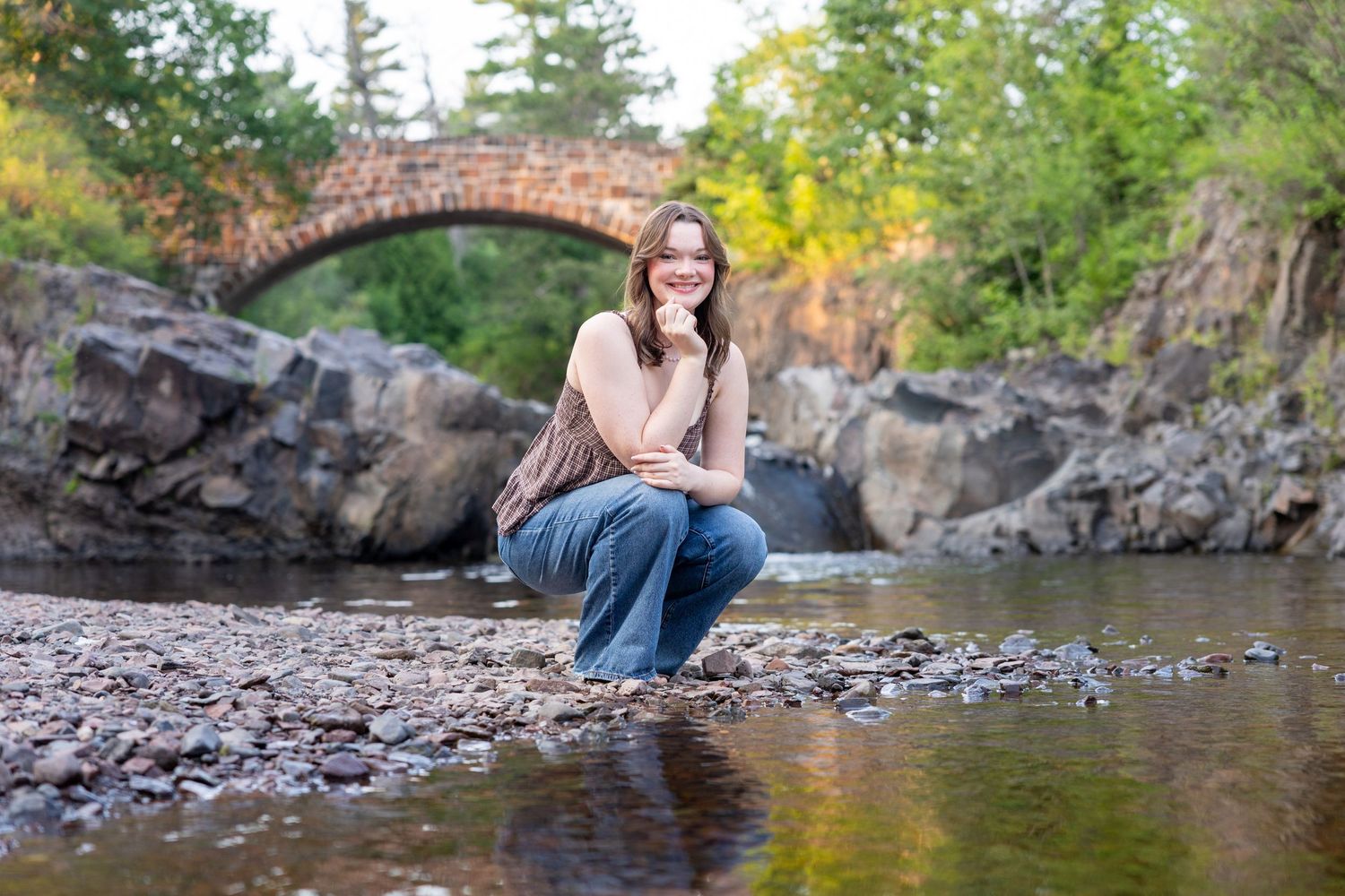 A young woman smiles while crouching by a rocky stream with a stone arch bridge in a lush forest background.