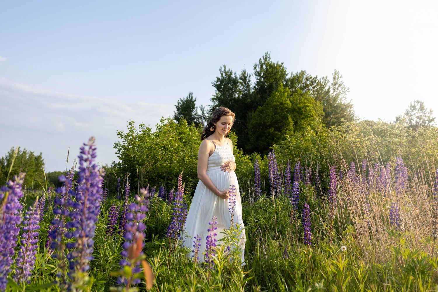 A person in a white dress poses among blooming purple lupine flowers in a sunlit meadow during golden hour.