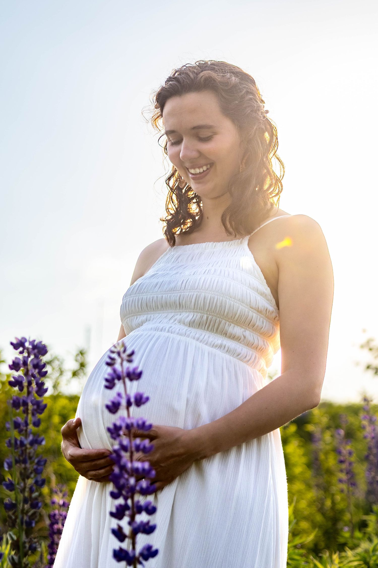 A pregnant person in a white dress stands in a field of purple lupine flowers at sunset during a maternity photoshoot.