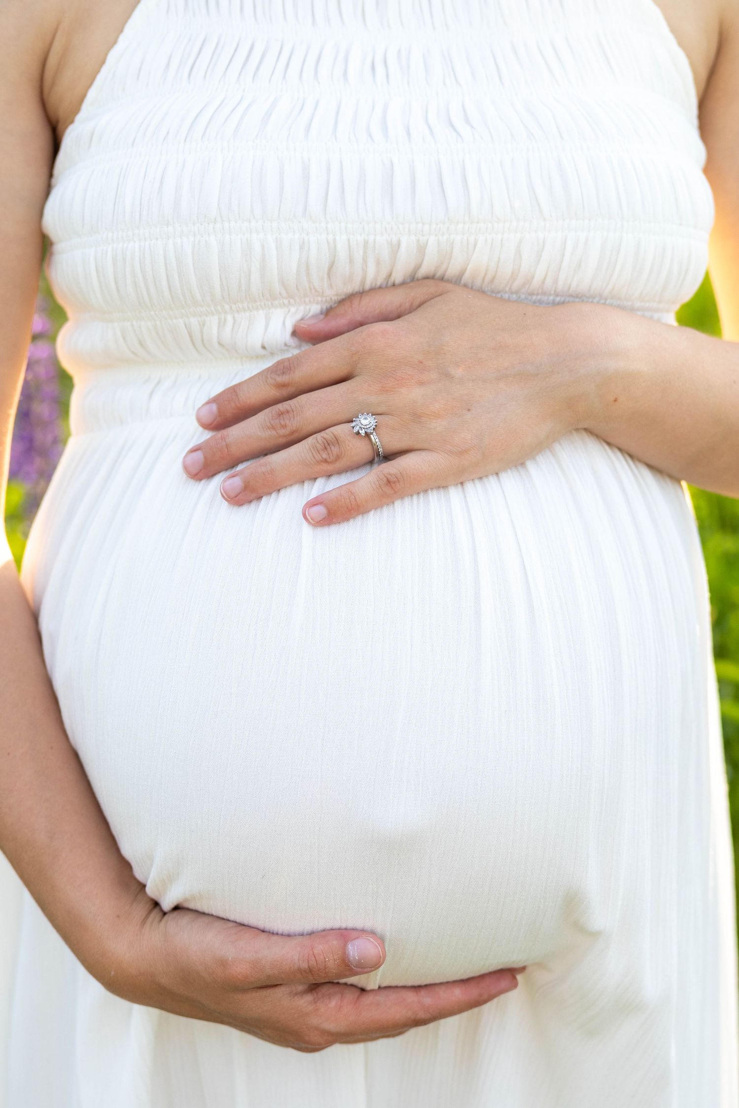 A pregnant woman in a white maternity dress holds her belly while showing off her engagement ring.