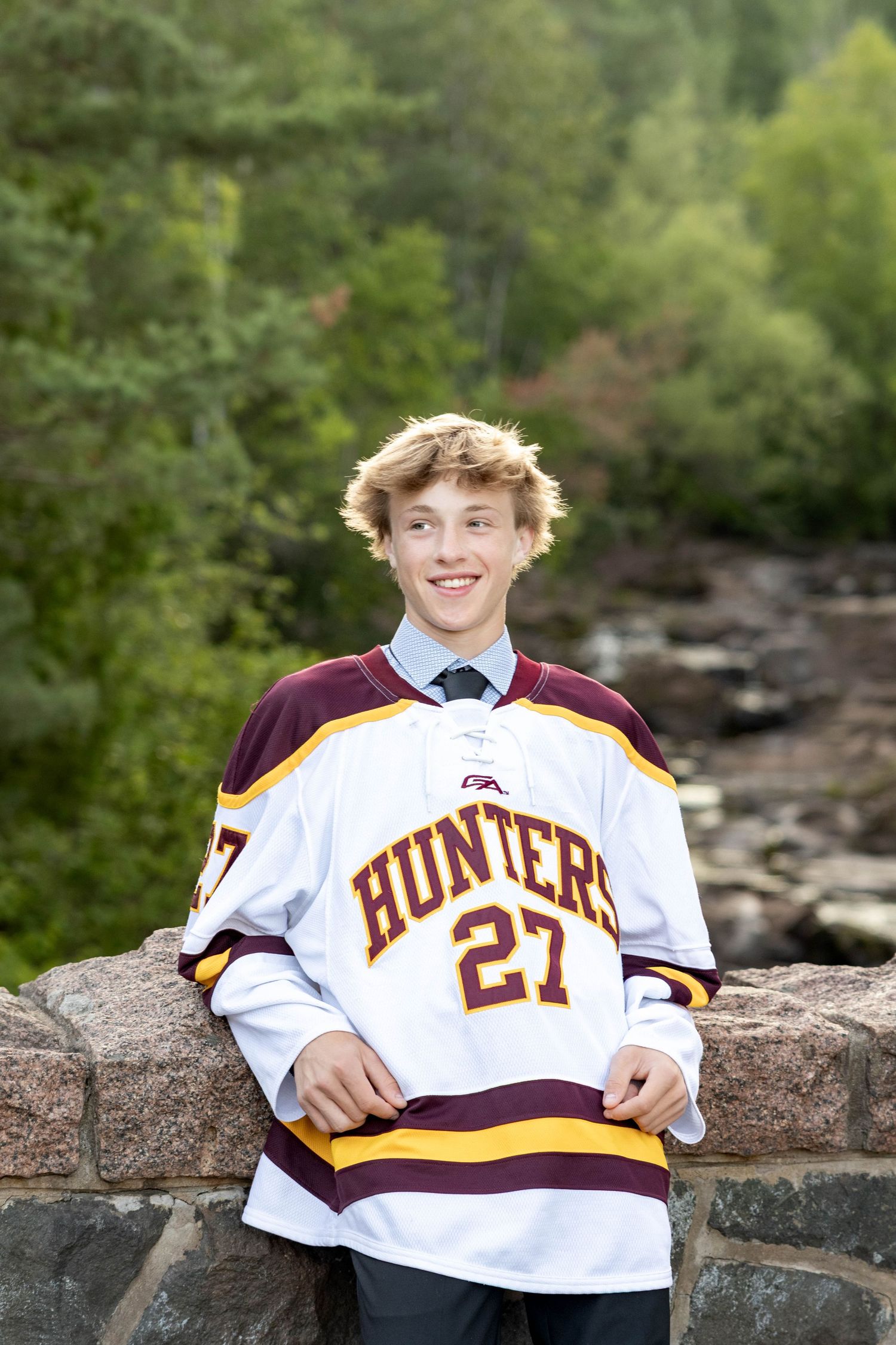 Student athlete poses on stone wall wearing white and maroon Hunters hockey jersey number 27 with scenic nature backdrop.