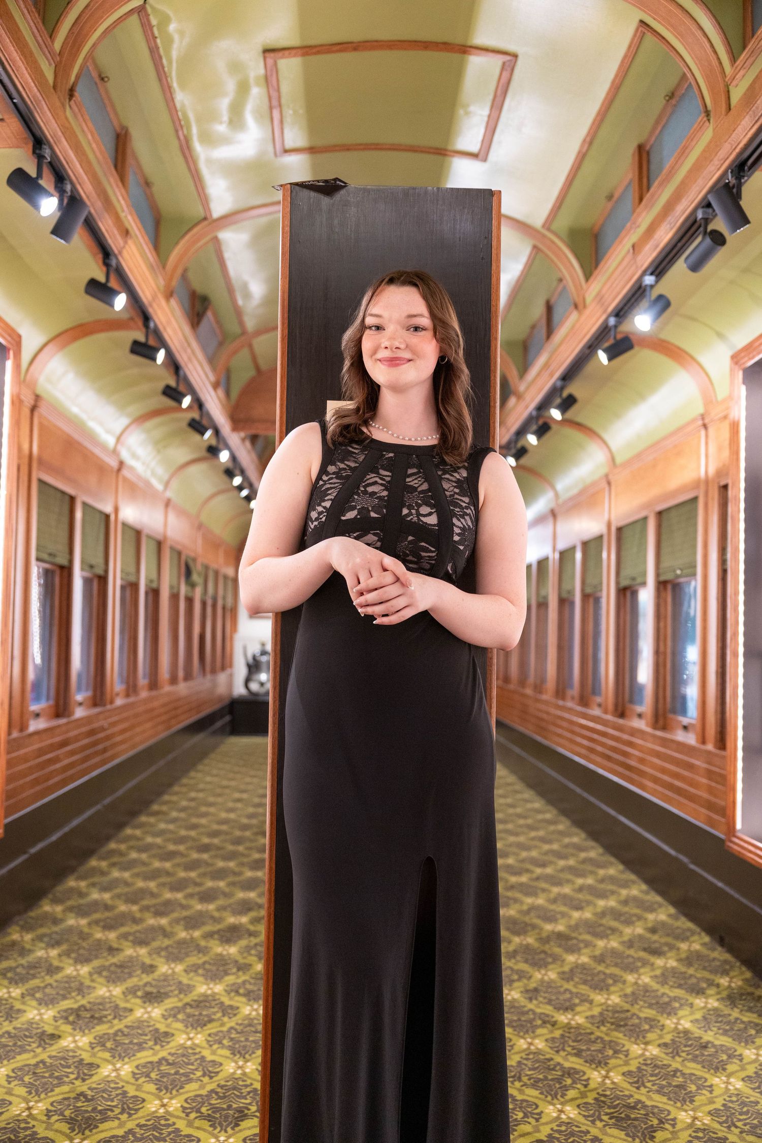 Young woman in elegant black gown poses inside a vintage train car with ornate wood paneling and green decor.