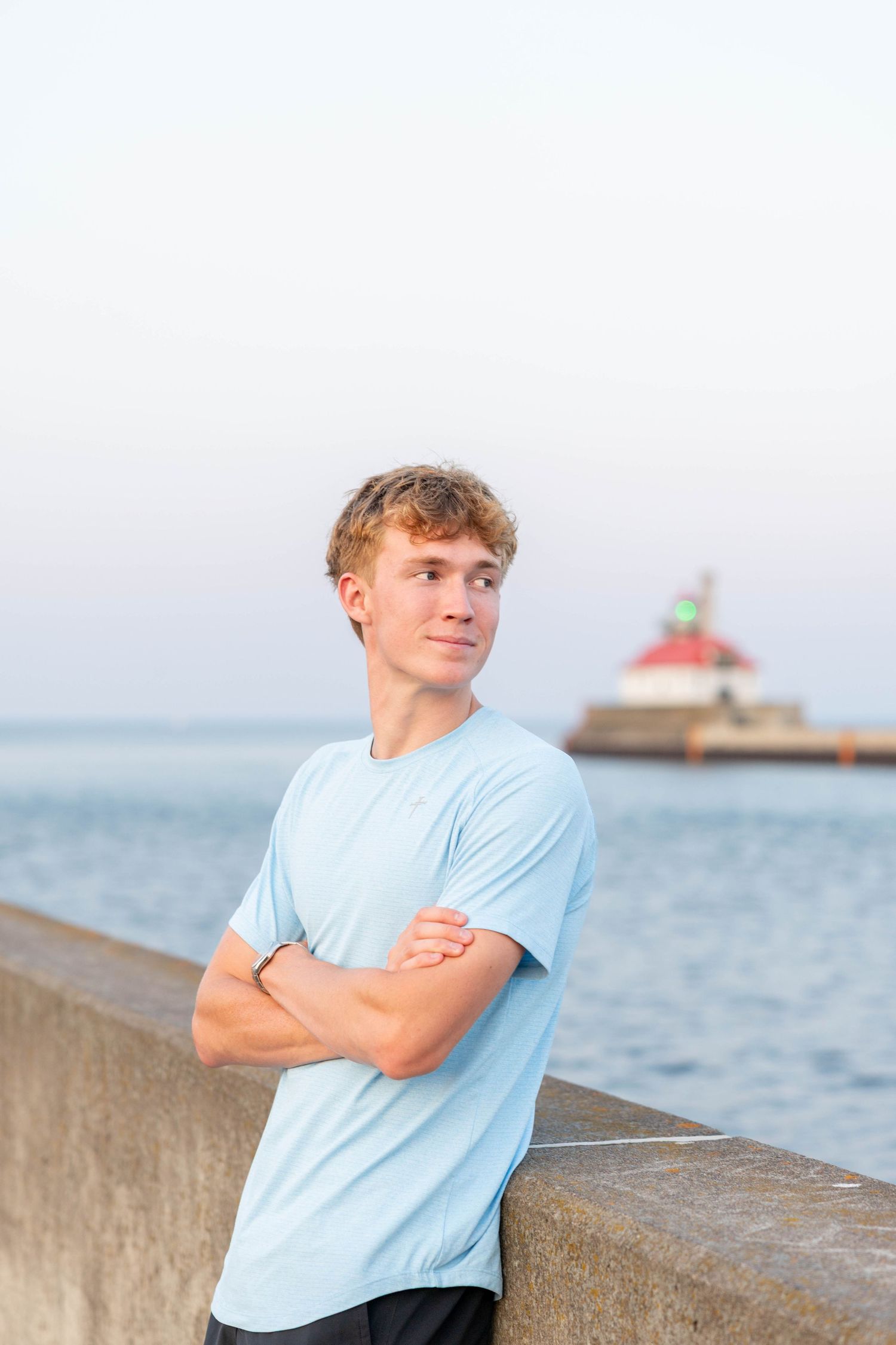 Young man in light blue t-shirt stands arms crossed by waterfront with red-roofed lighthouse in background.