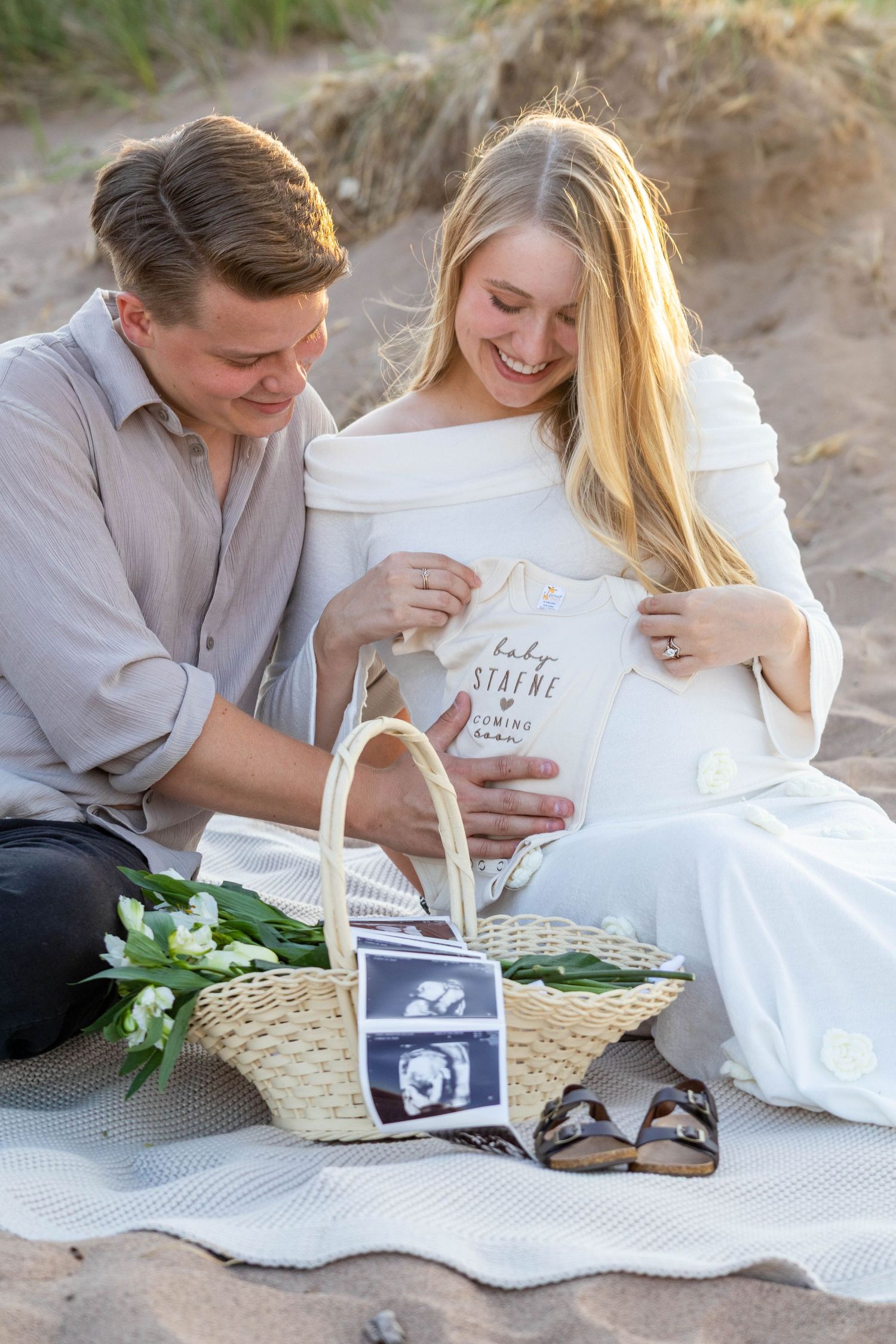 A couple lying on a beach blanket shares a tender moment while looking at ultrasound photos and white flowers.