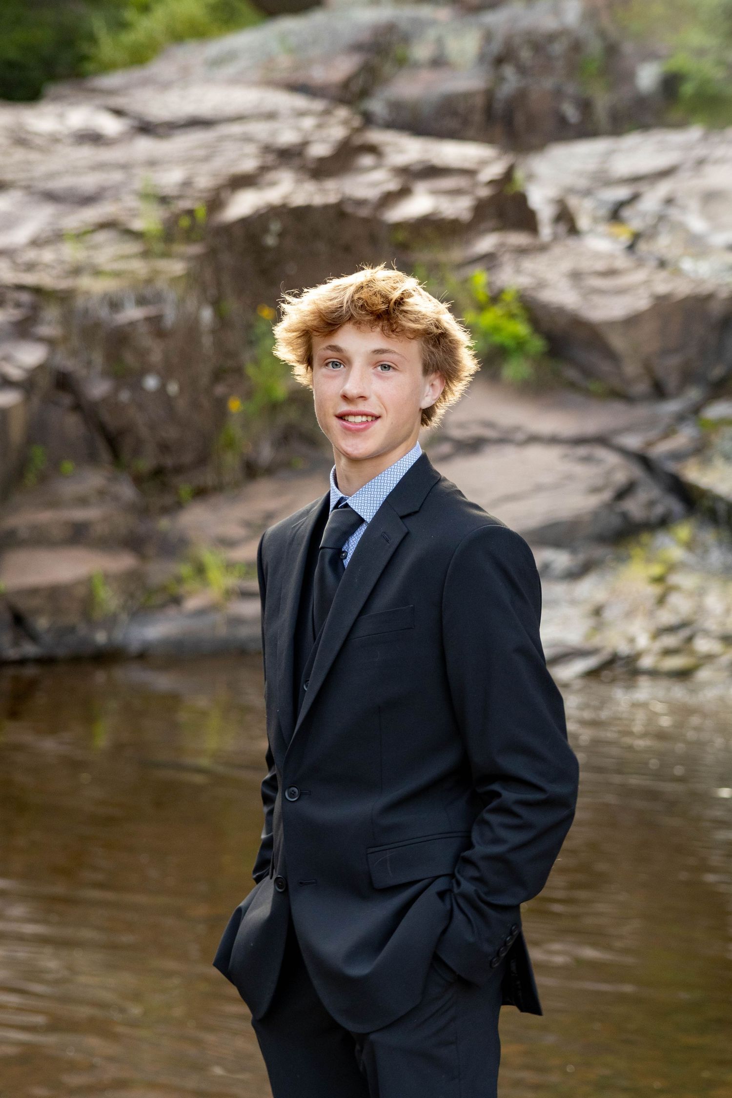 A person in a black suit poses outdoors near a rocky riverbank with water and natural scenery in the background.