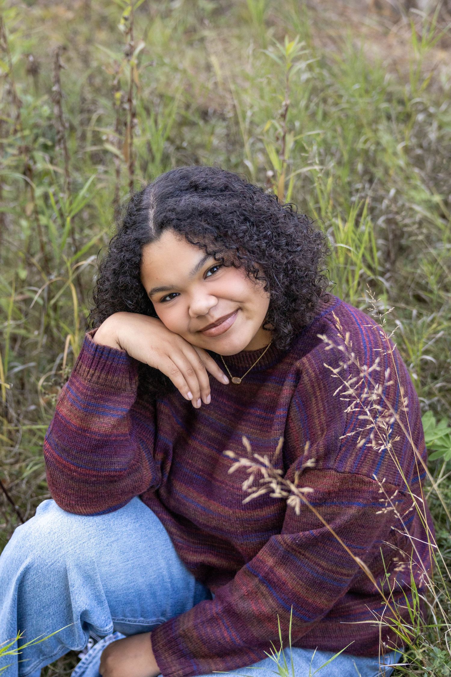 Someone wearing a purple striped sweater sits outdoors in jeans among tall grass on an autumn day.
