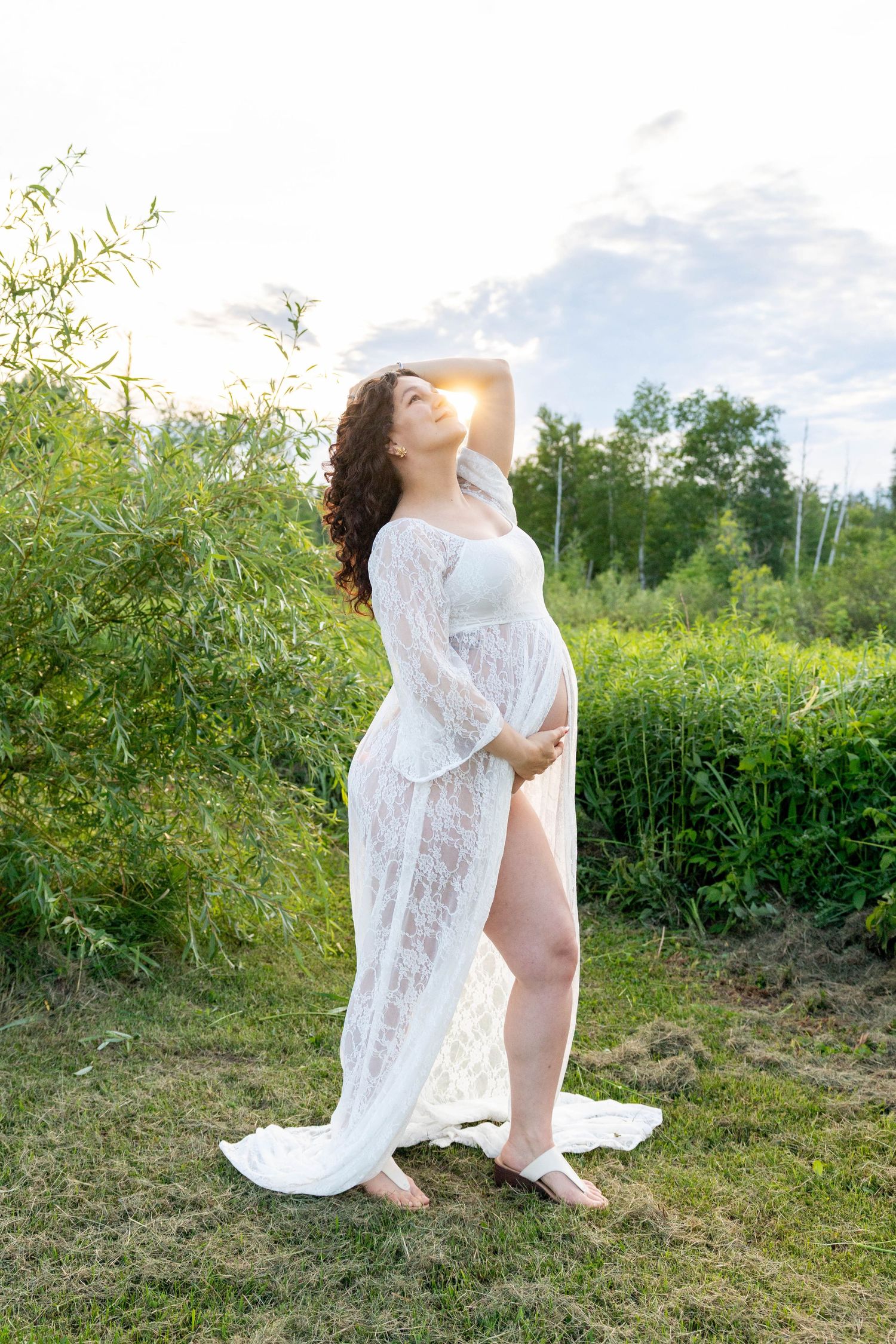 Pregnant woman in white lace maternity gown poses outdoors in lush green nature setting during golden hour sunset.