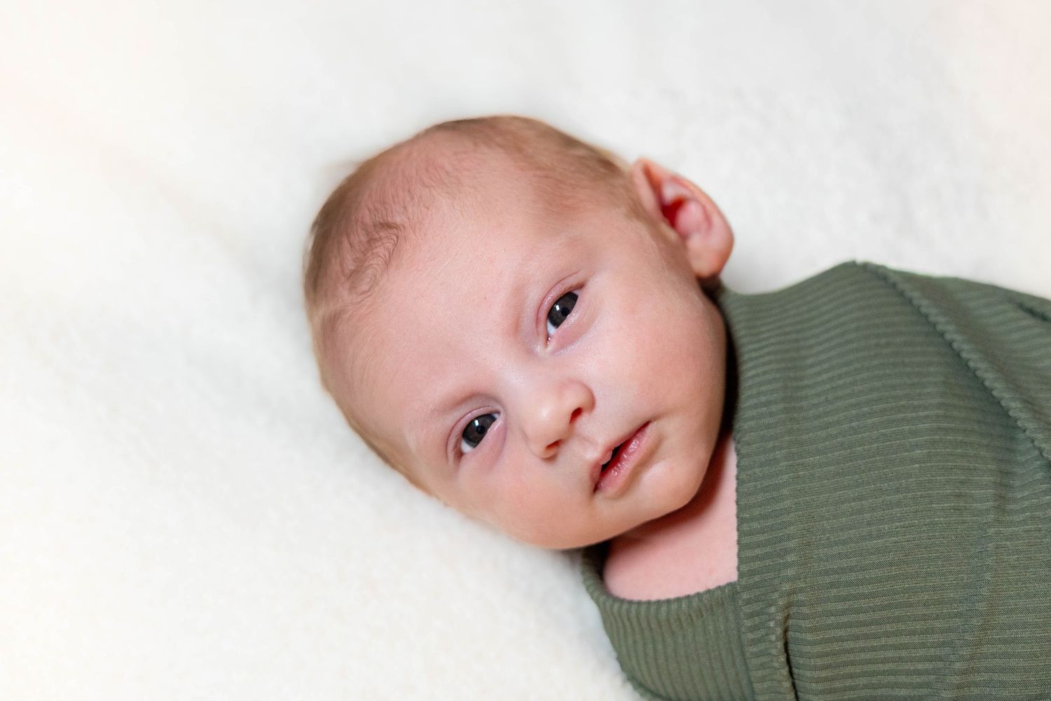 Baby wearing green shirt lies on white blanket looking at camera with curious expression.