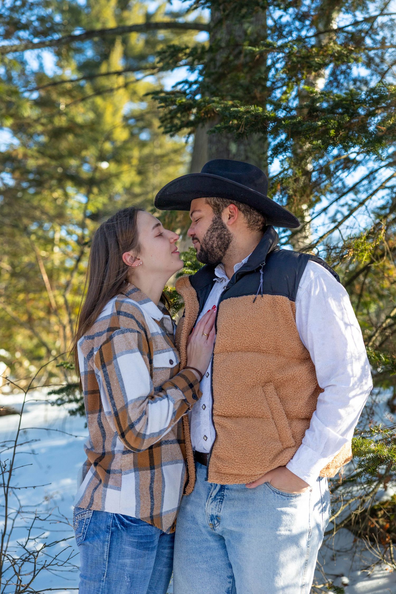 Romantic winter engagement photos show couple in matching brown vests sharing intimate moments in snowy forest setting.