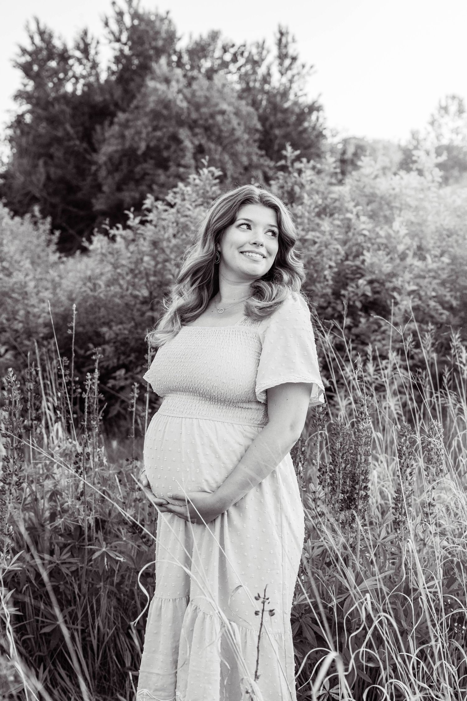 A woman in a white dress poses gracefully in a natural field setting with tall grass during a monochrome photoshoot.