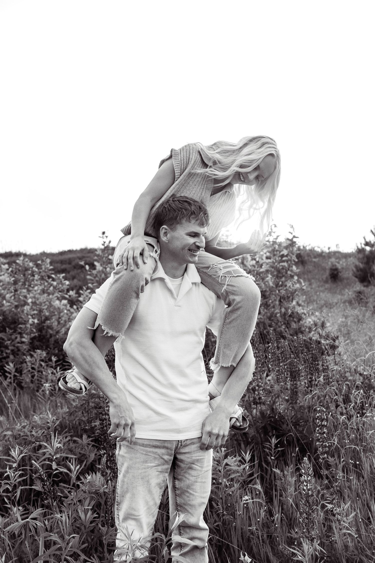 A black and white photo of a couple playfully posing in a field with tall grass and foliage in the background.