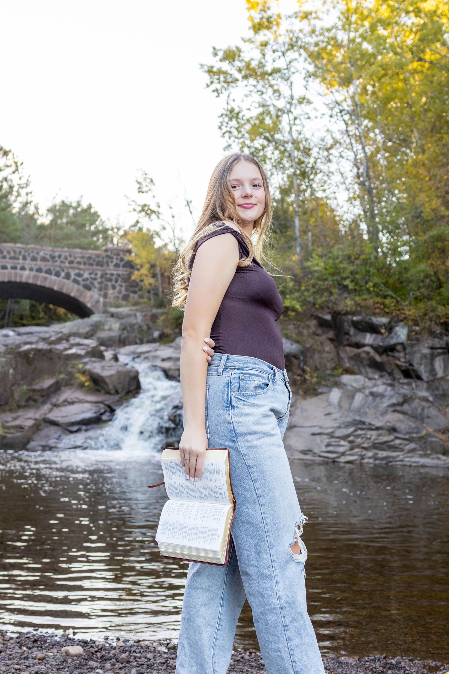 Person in light denim overalls and brown top poses near a scenic waterfall and stone bridge on an autumn day.