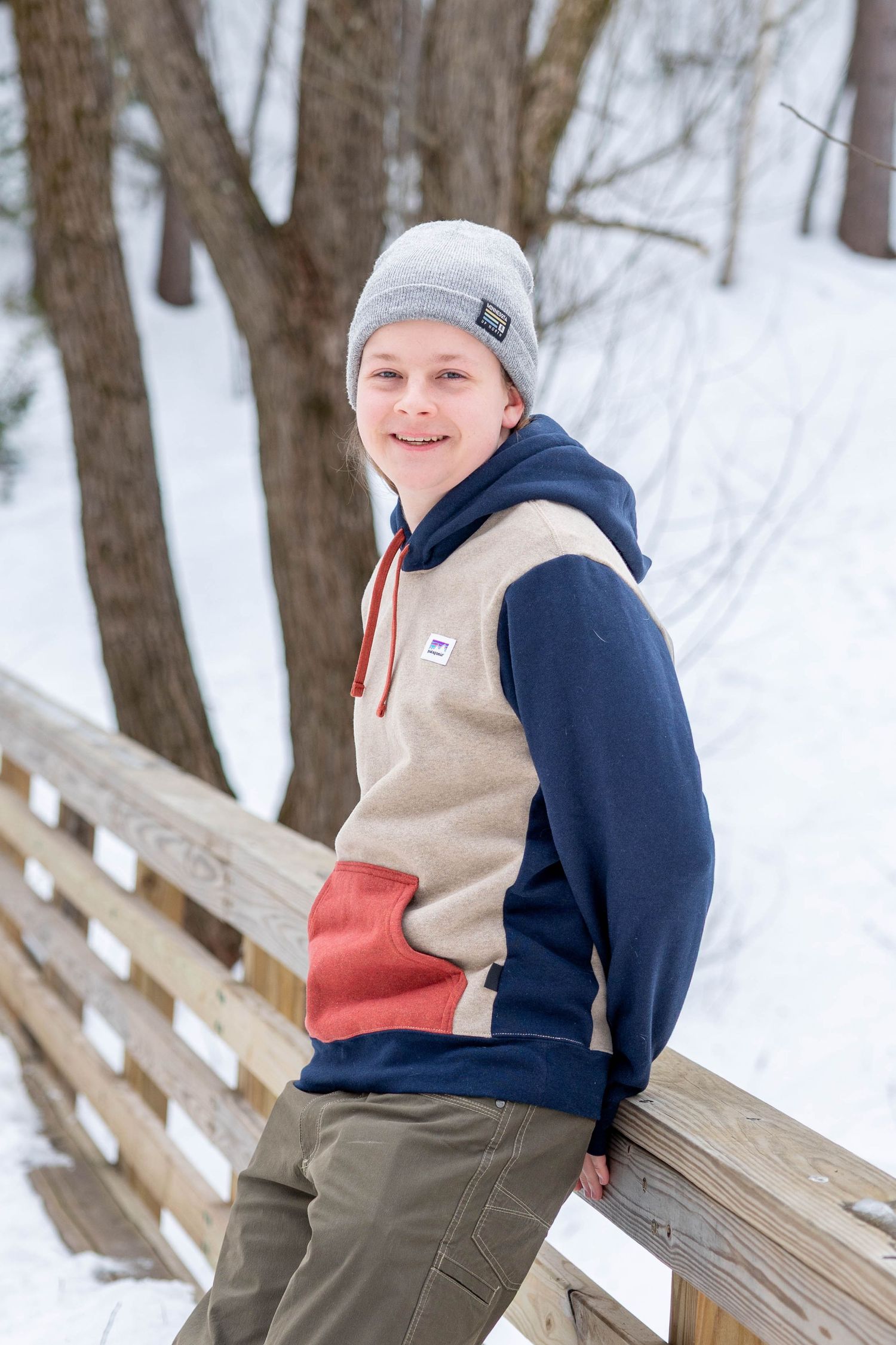 Smiling teenage boy in colorful hoodie and gray beanie leaning on wooden railing in snowy winter park.