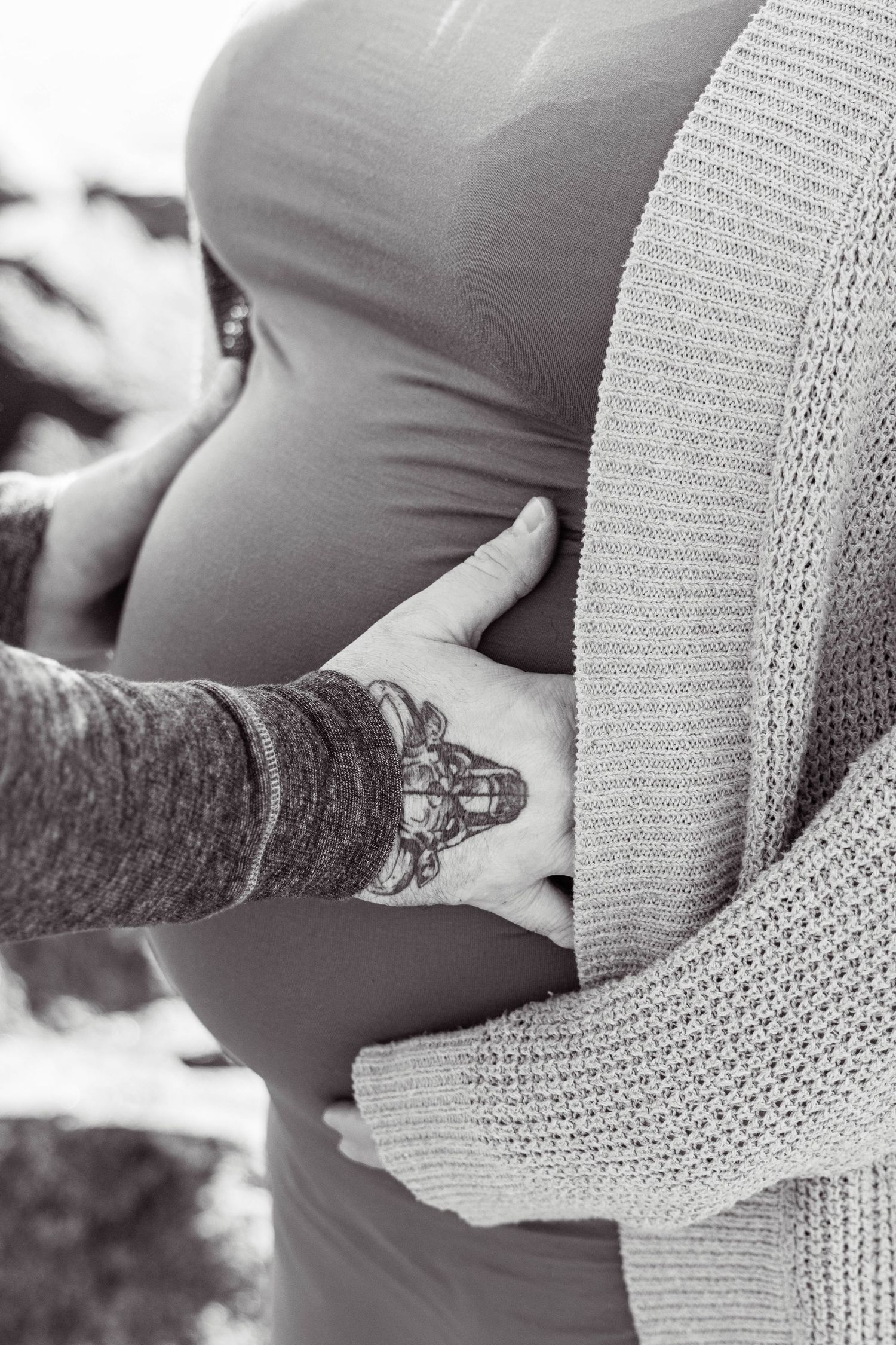 A tattooed arm gently touches a pregnant belly against a soft knit sweater.