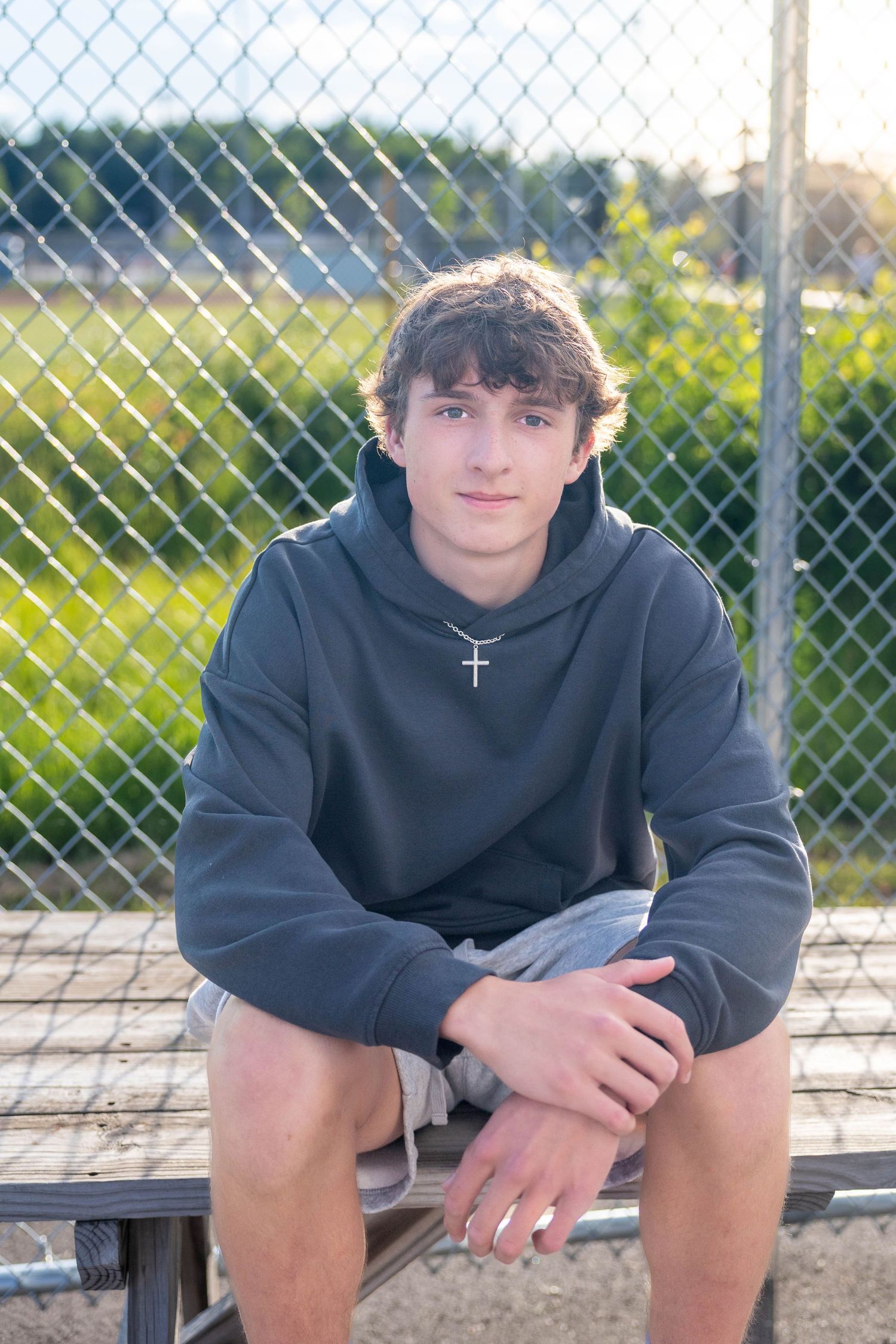 A teenage boy in a gray hoodie with a cross necklace sits on bleachers by a chain-link fence outdoors.