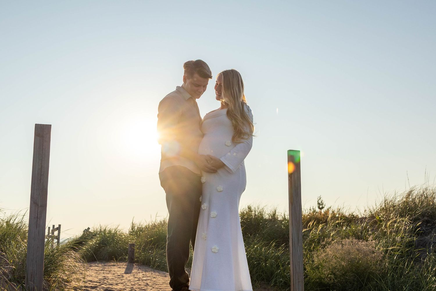 Silhouetted couple embraces in a romantic pose against a bright sunset backdrop with tall grass and fence posts.