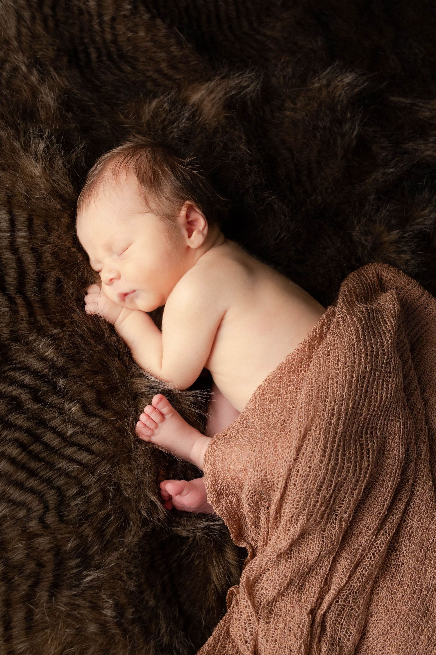 Sleeping newborn partially wrapped in brown blanket on textured fur background.