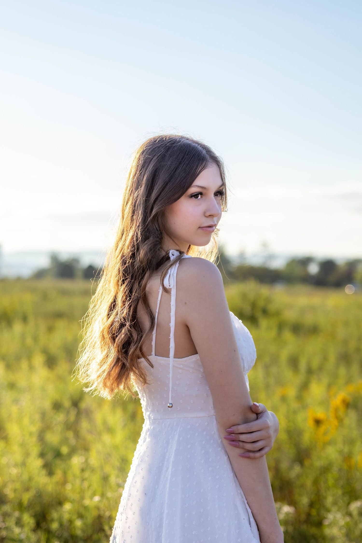 A woman in a white dress poses in a sunlit field of wildflowers during golden hour.