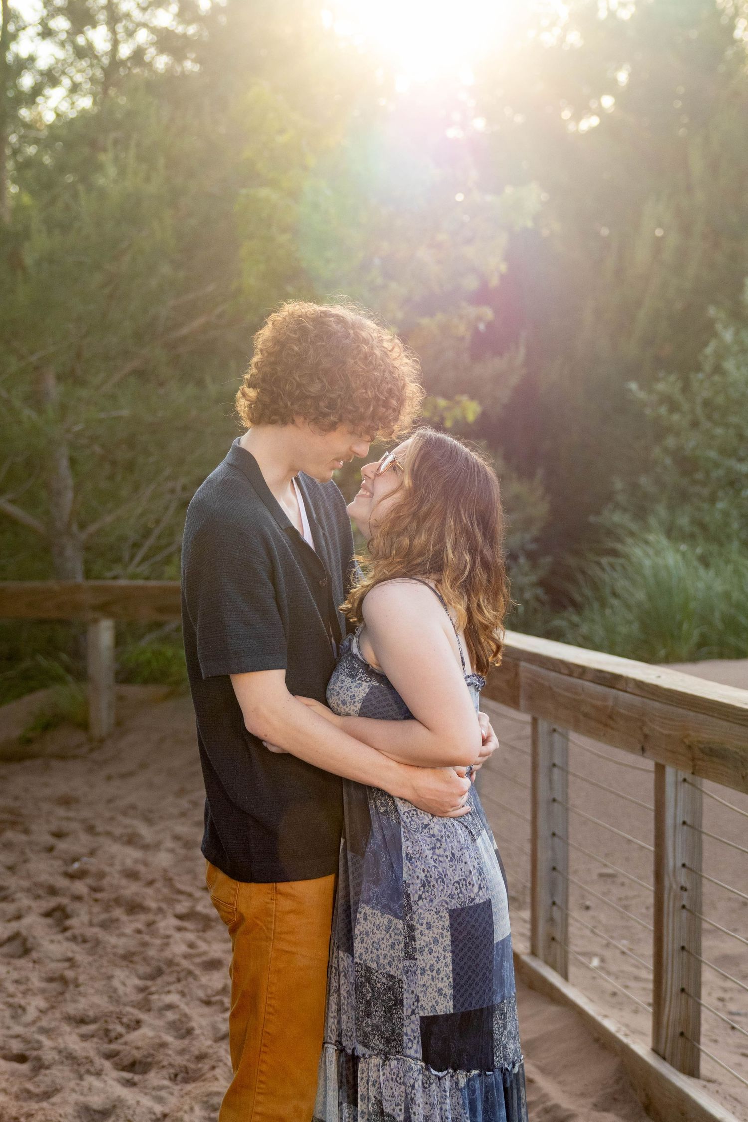 A romantic couple embraces on a wooden boardwalk during golden hour with sunlight streaming through the trees.