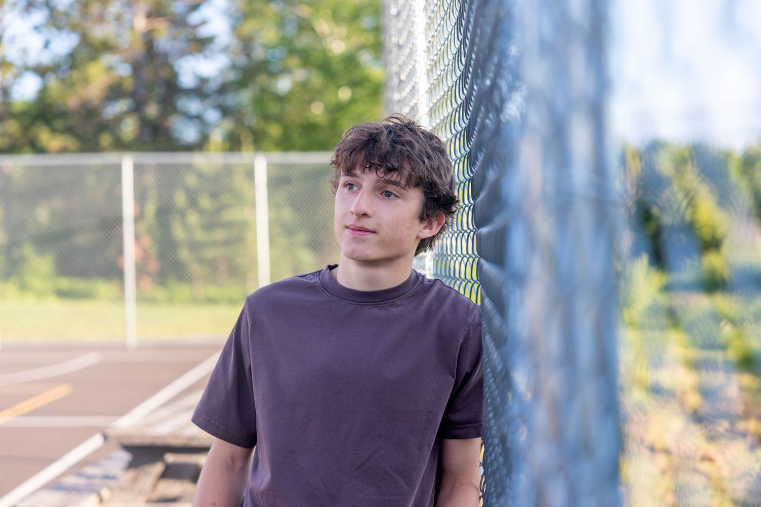 Young man in brown t-shirt leaning against chain-link fence at tennis court, looking thoughtfully into distance.