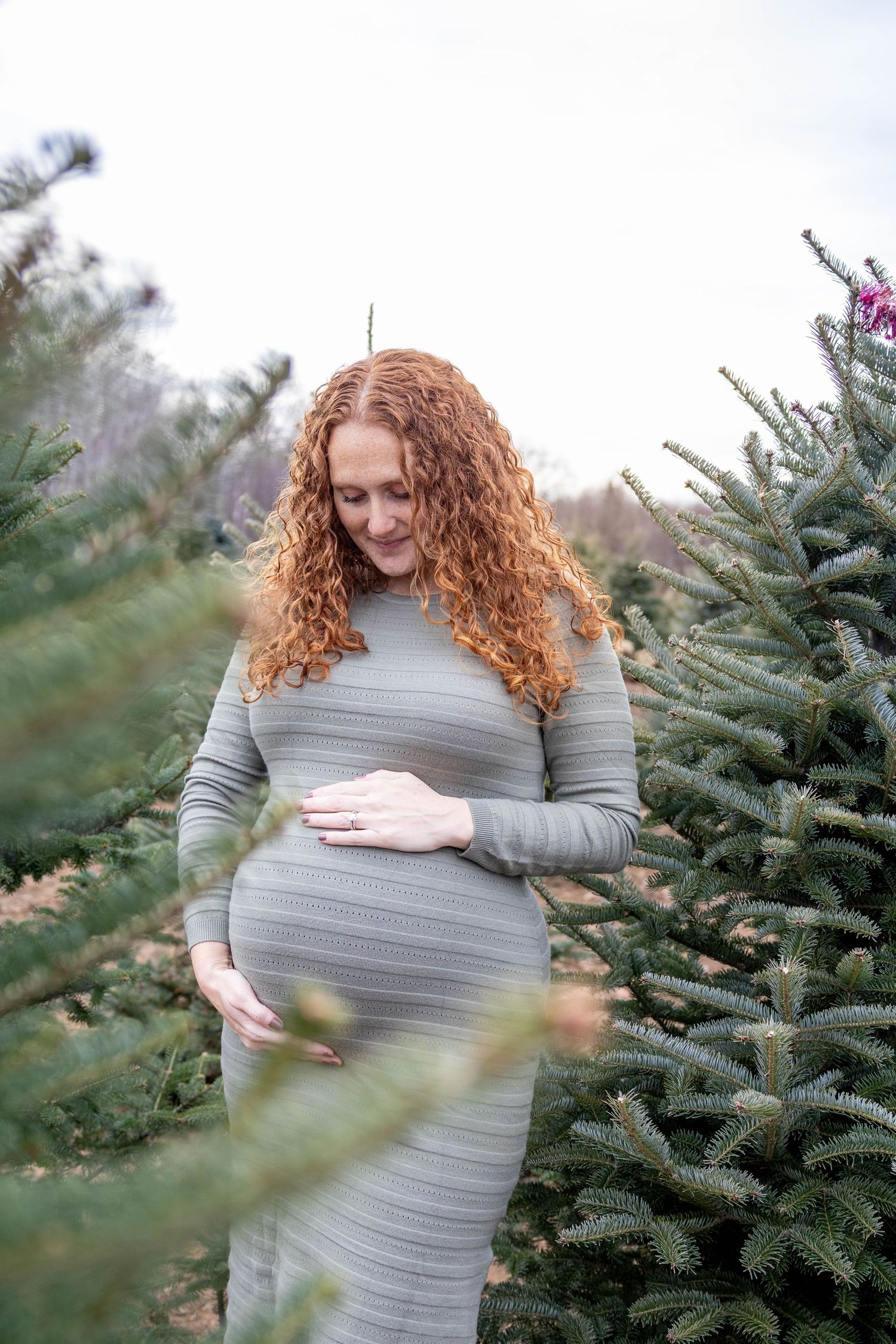 Expecting mother in a grey striped dress poses among evergreen trees during a winter maternity photoshoot.