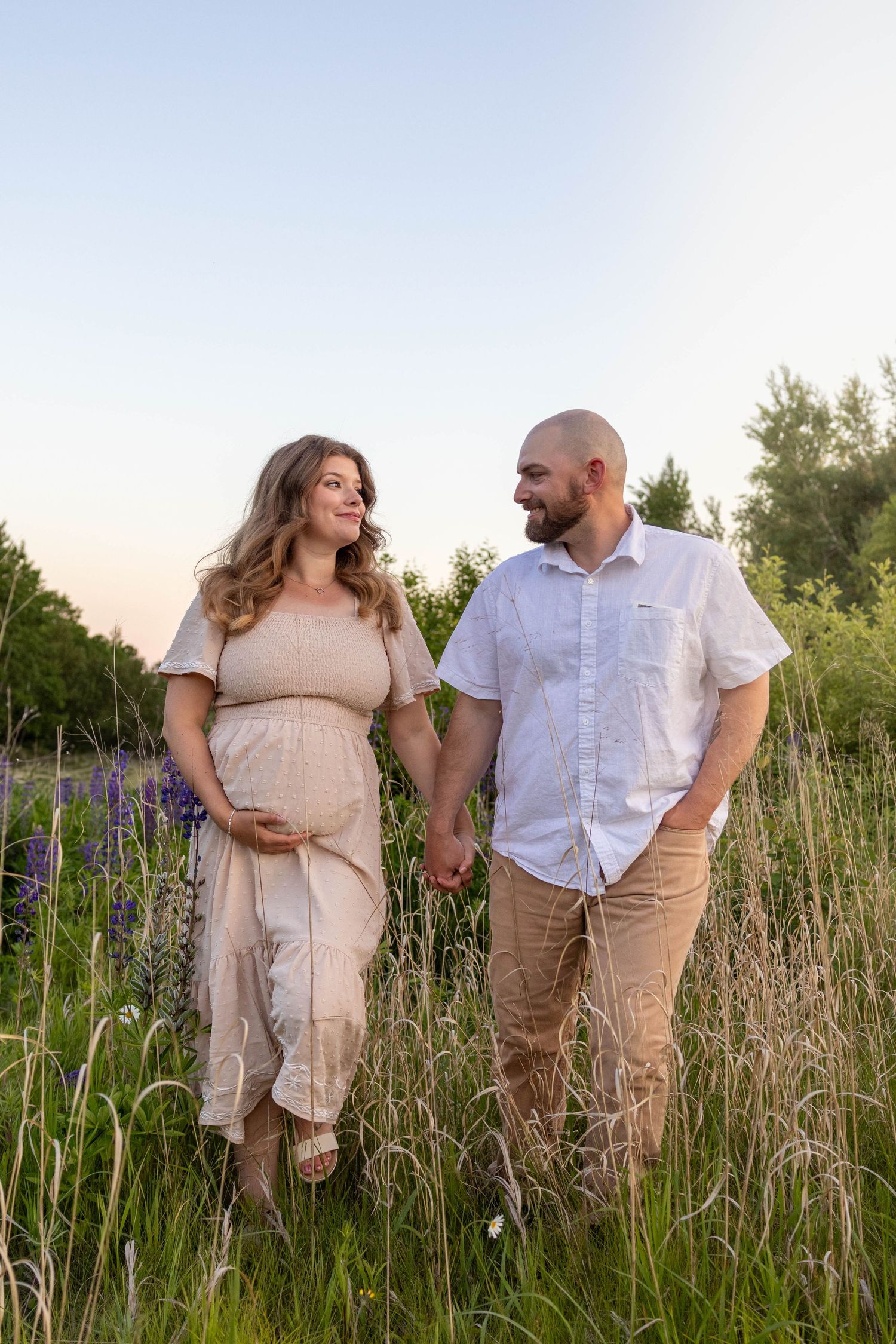 Couple stands together lovingly in a tall grass field at sunset with rustic neutral outfits.