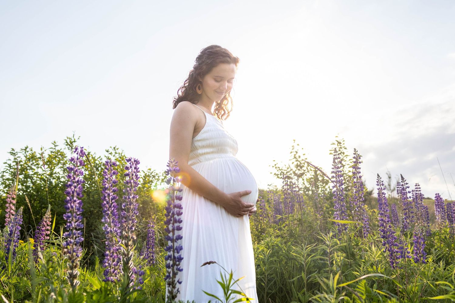 Person in a white dress stands among vibrant purple lupine flowers during golden hour at sunset.