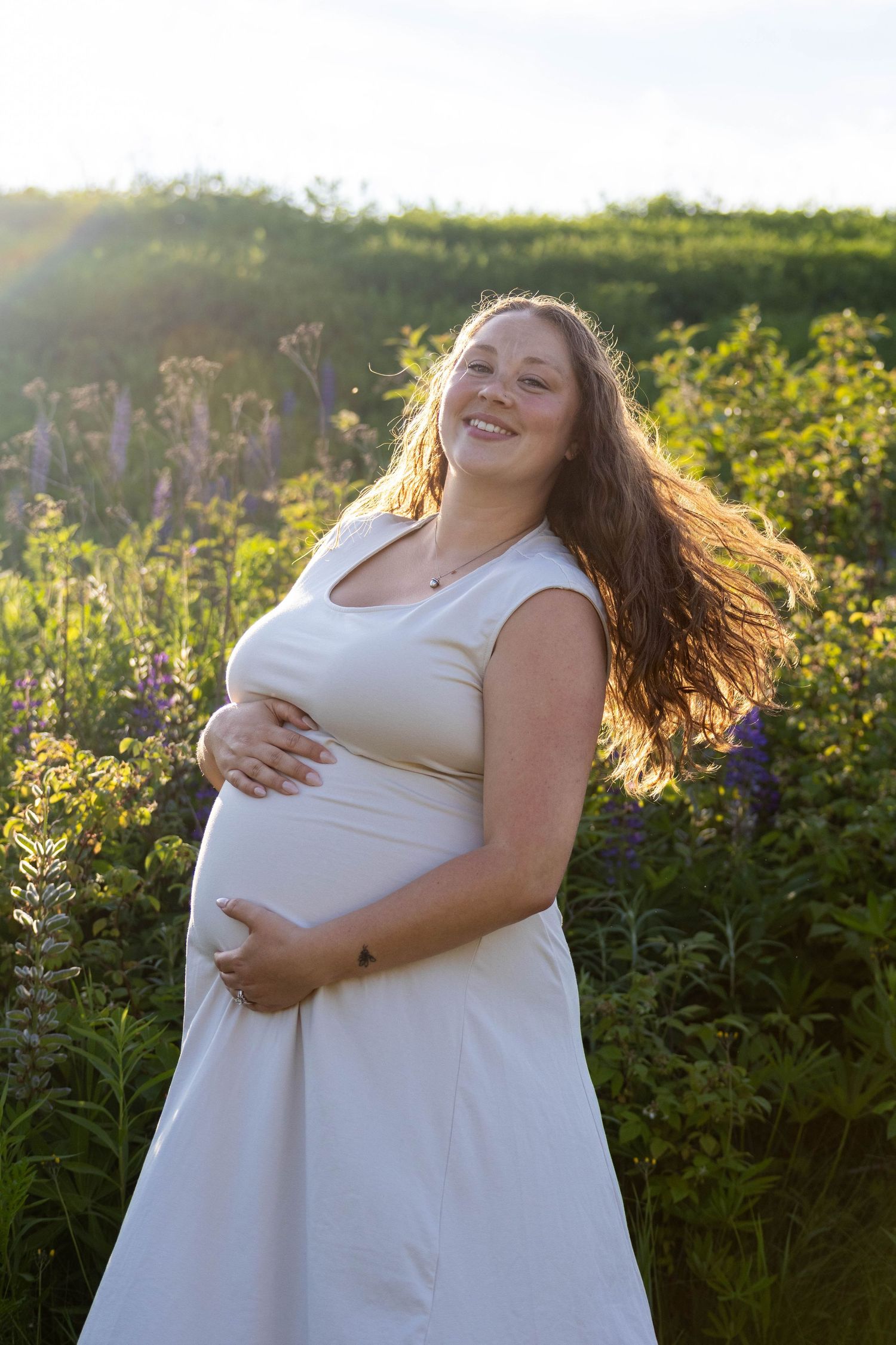 A person in a white dress cradles their pregnant belly while standing in a sunlit field of yellow flowers at golden hour.