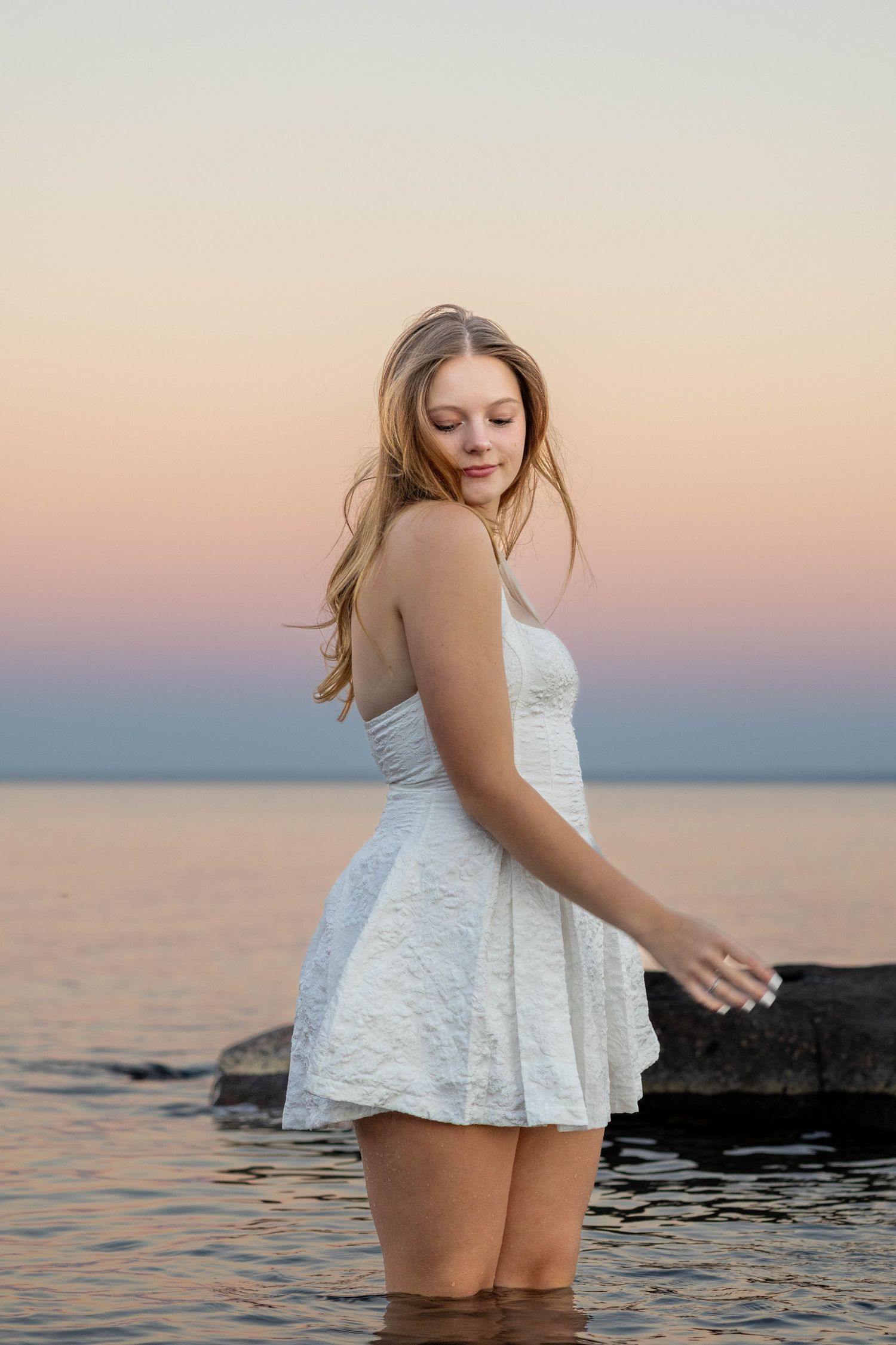 Woman in white lace dress poses in water at sunset with pink and purple sky reflecting on ocean surface.