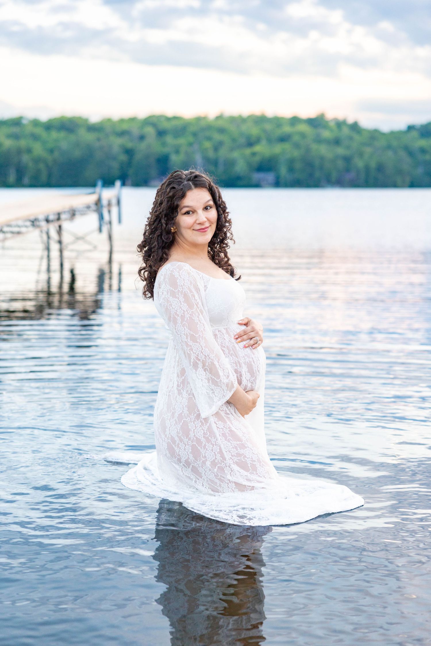 Pregnant woman in white lace dress standing in calm lake water, smiling, with a dock and green trees in background.