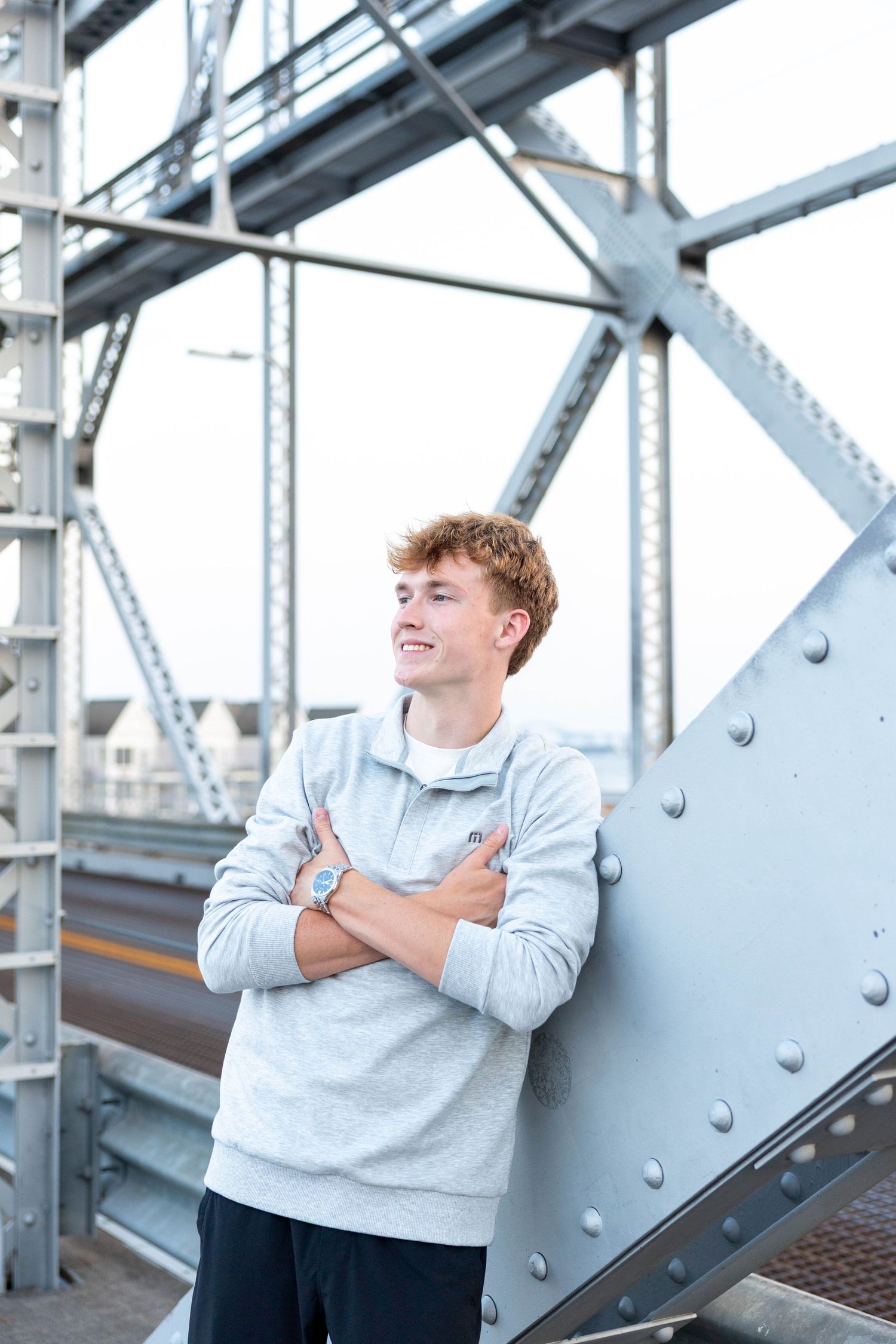 Young man with red hair in gray sweatshirt stands arms crossed on steel bridge structure, smiling confidently.