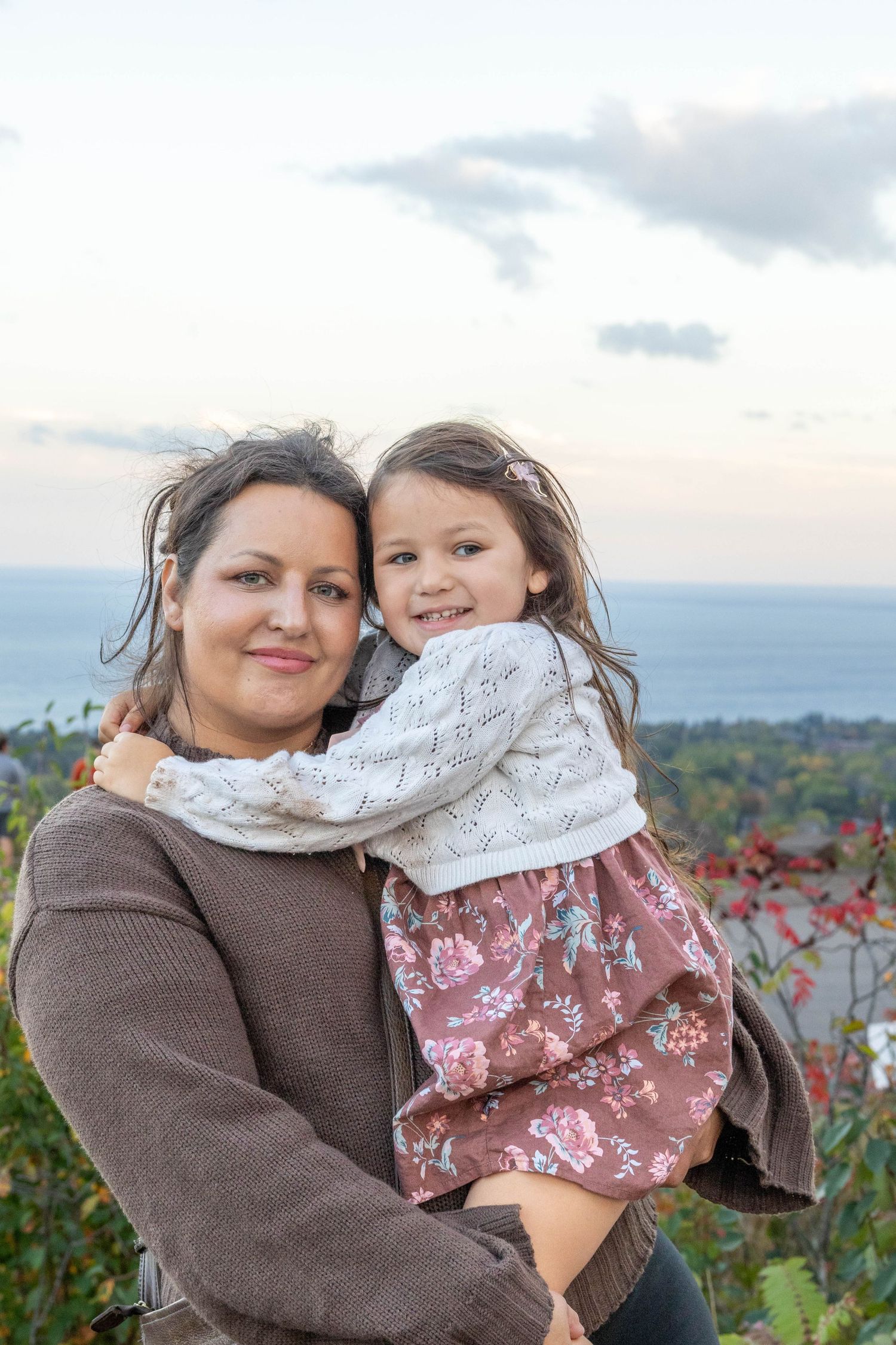 A parent and child embrace outdoors against an autumn landscape with colorful fall foliage in the background.
