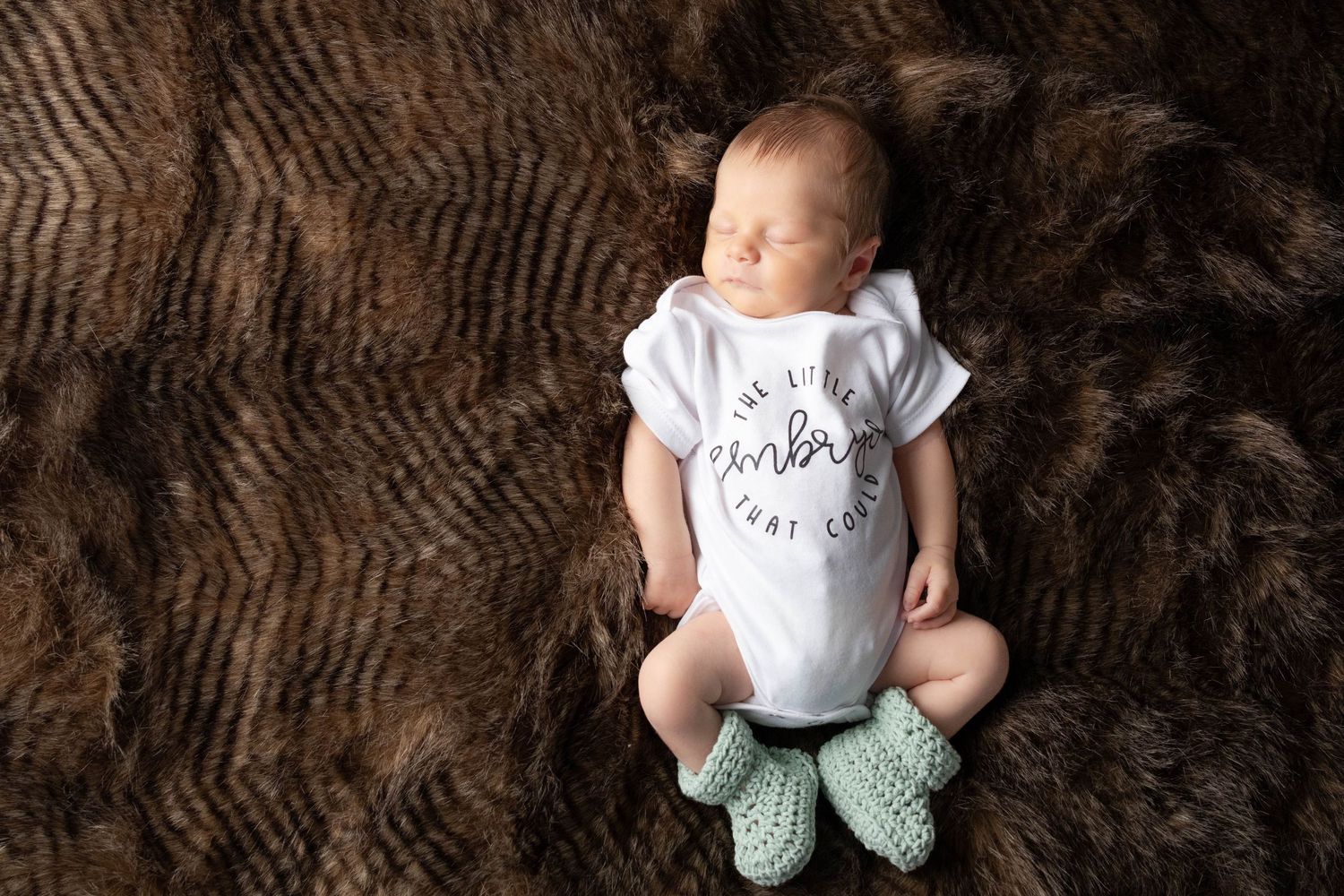 Baby wearing white onesie and mint booties lies on textured brown backdrop.