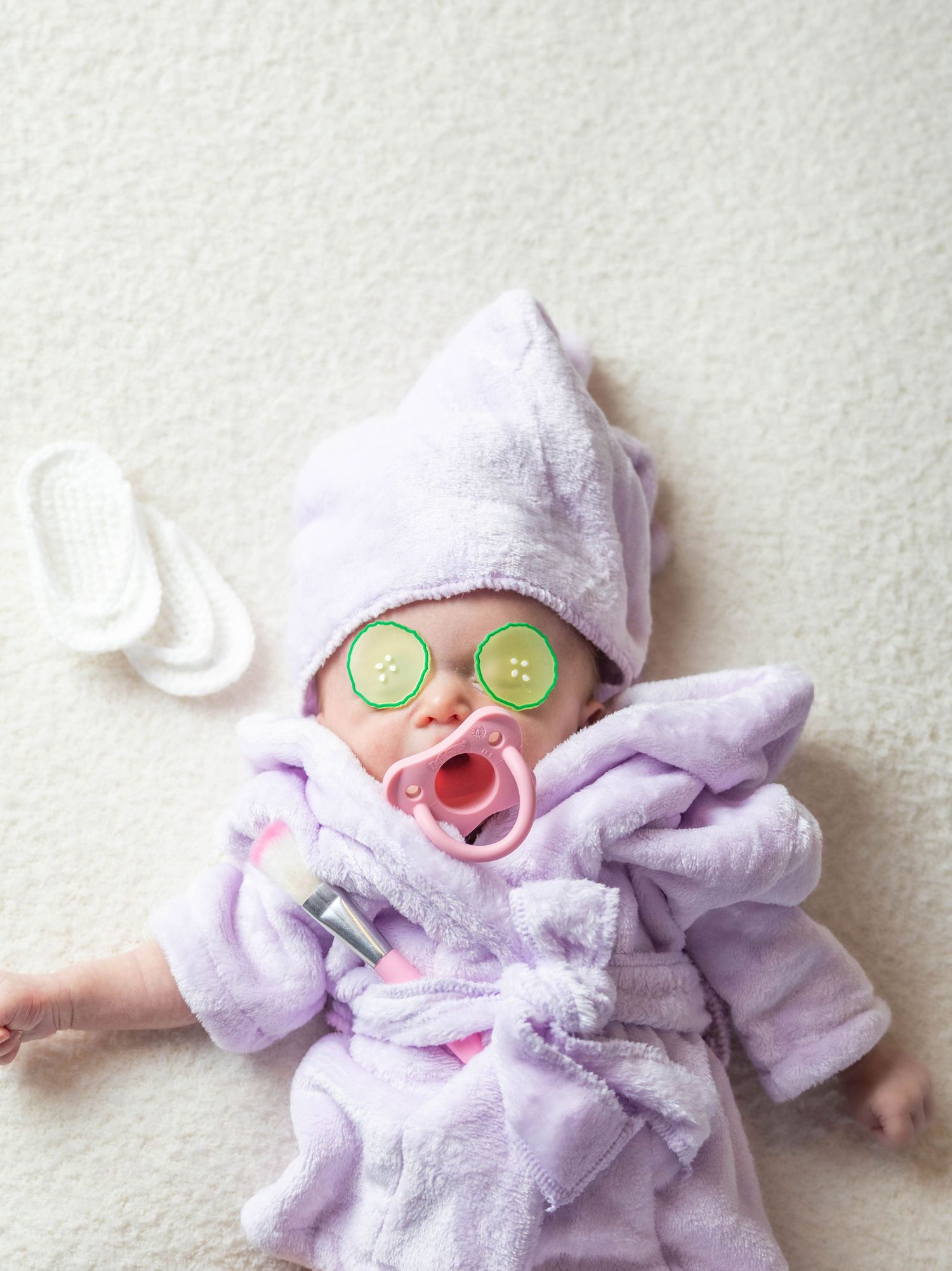 Playful baby in purple robe and cucumber spa mask relaxing on white background.