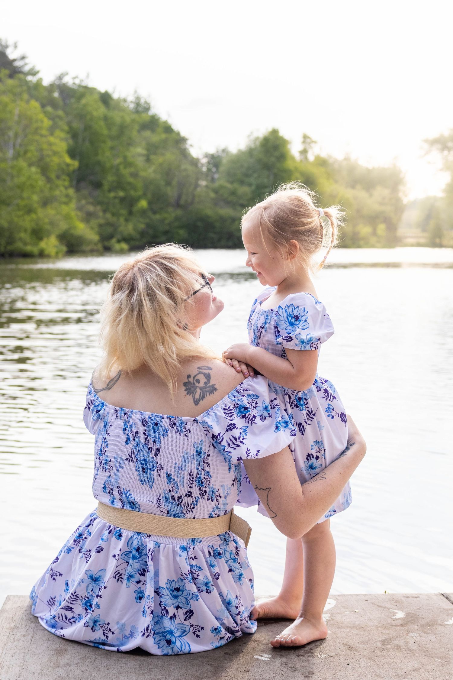 Two people in matching blue and white floral dresses sit by a serene lake at sunset.