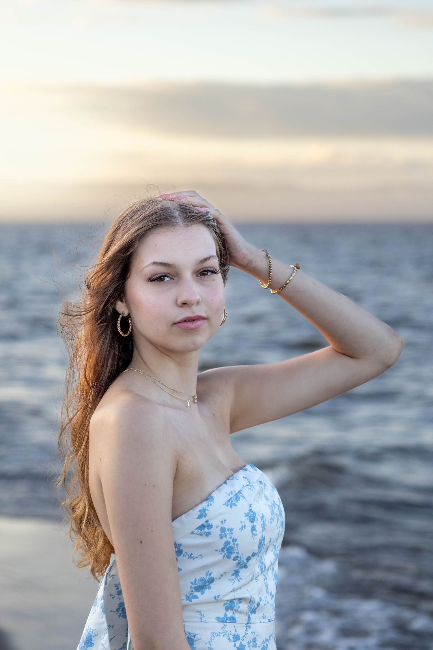 A person in a white floral dress poses by the ocean at sunset.