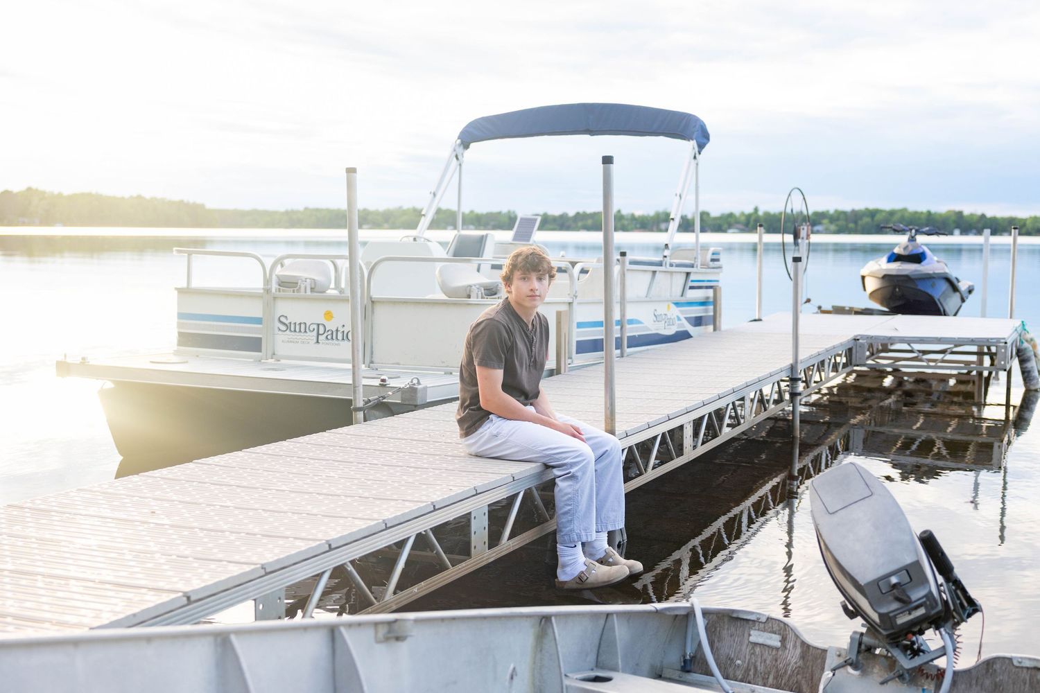 Young man sitting on a boat dock by a calm lake with a pontoon boat moored nearby at sunset.