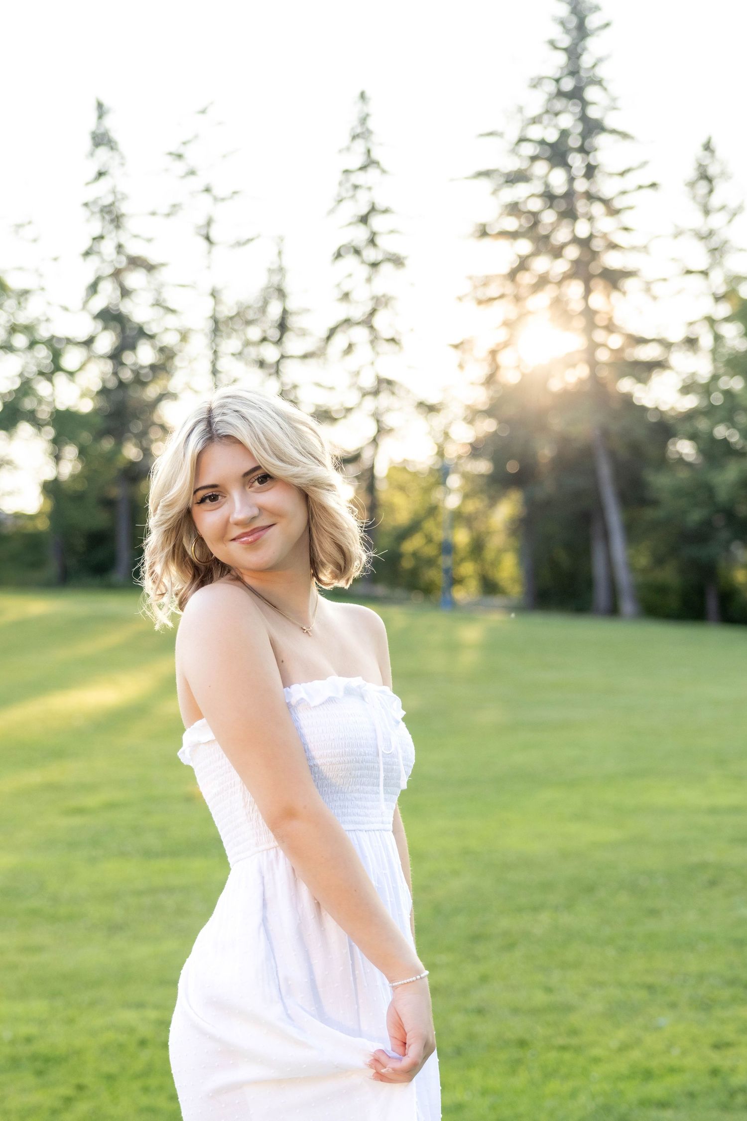 A woman in a white sundress poses in a grassy field with evergreen trees and golden sunlight in the background.