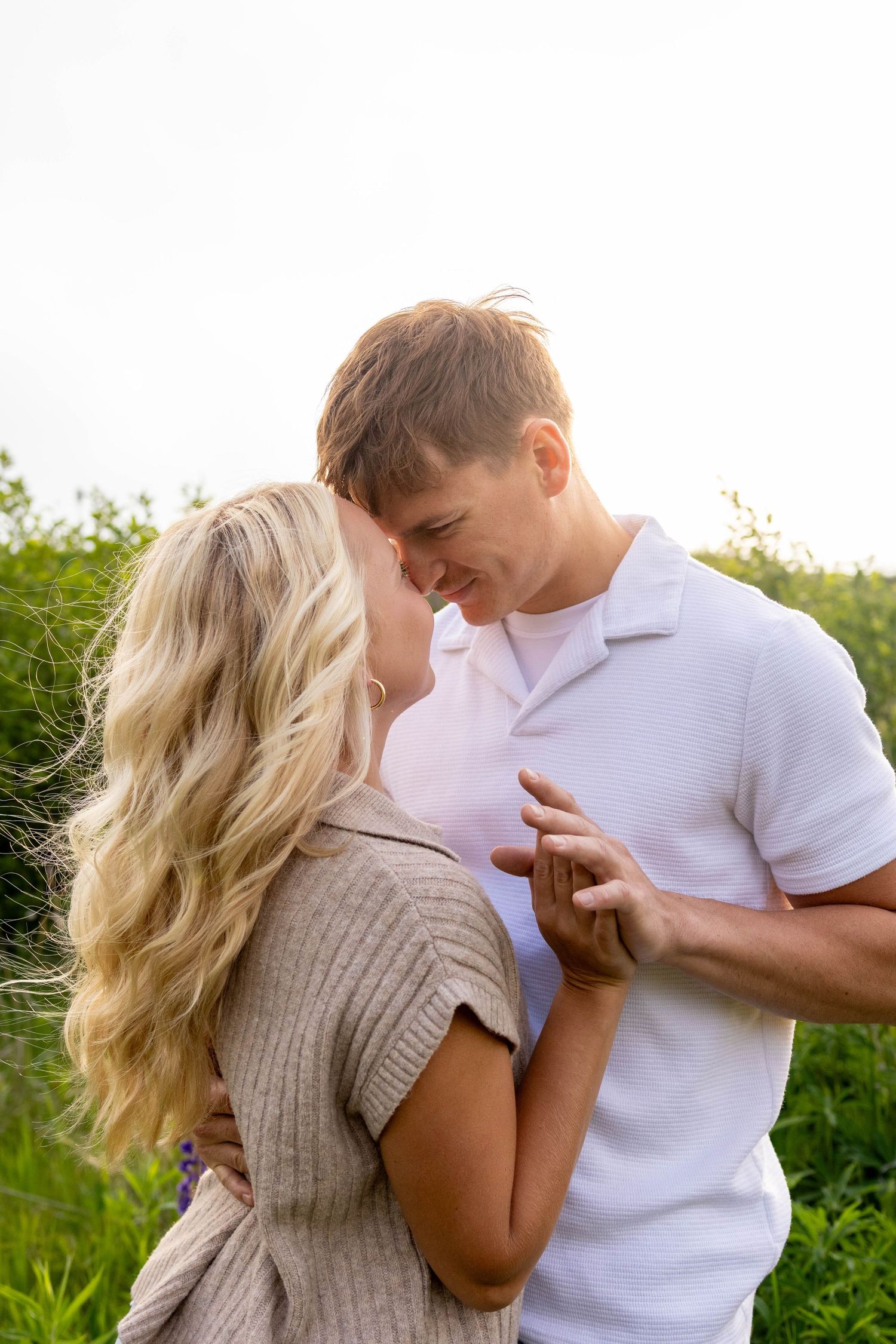 Romantic couple in white and beige clothing embraces intimately in a lush green outdoor setting.