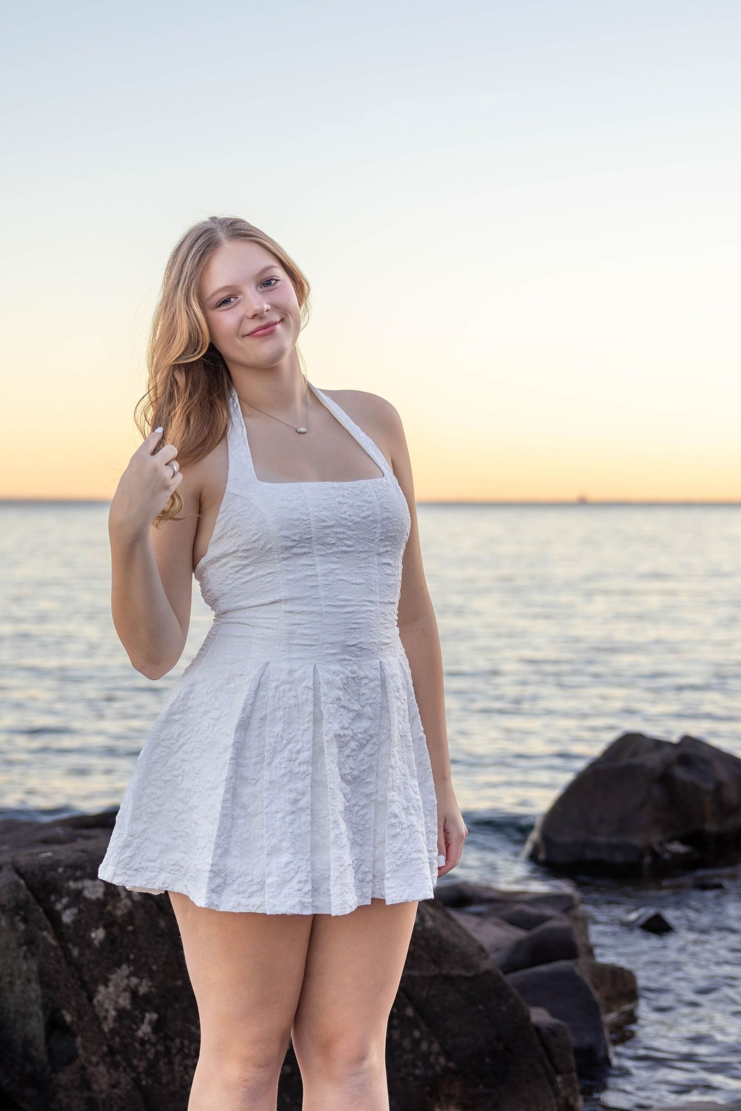 A person in a white lace dress poses on rocky beach during sunset near the ocean.