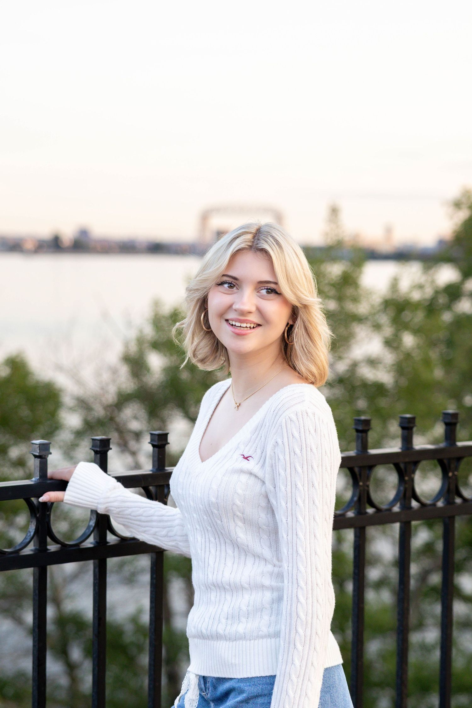 A person in a white cable knit sweater poses casually by a black iron railing with a waterfront view.