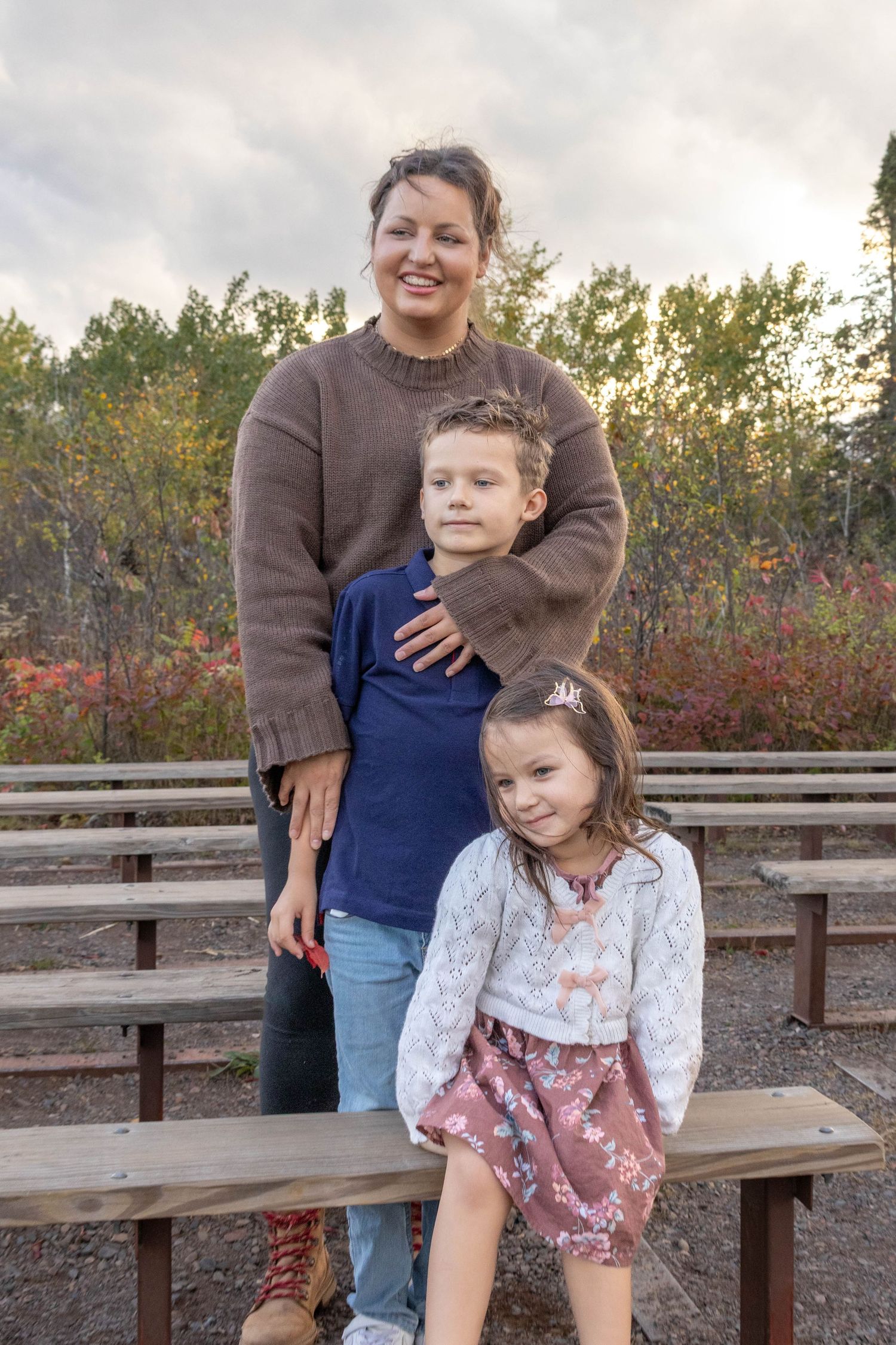 Family sitting together on wooden benches outdoors with autumn foliage in background.