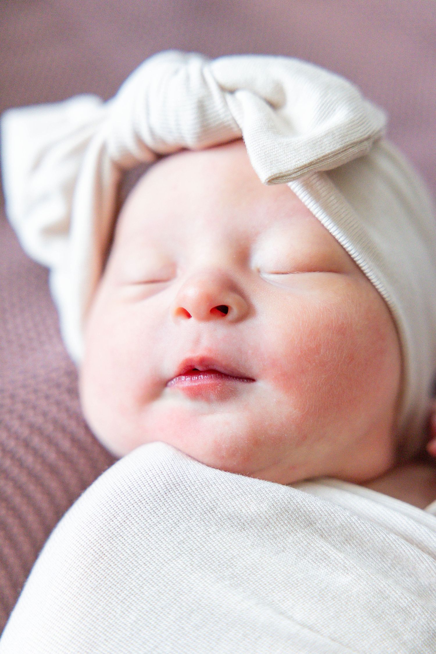 Sleeping baby wearing white headband bow rests peacefully on textured mauve surface.