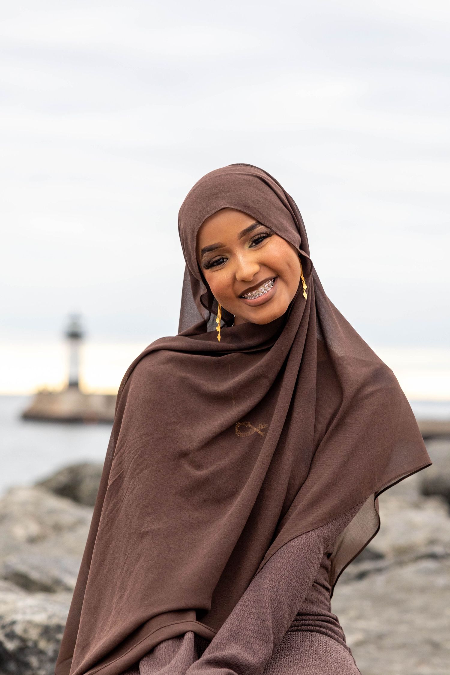 A person in a brown hijab poses outdoors near a lighthouse on a cloudy day.