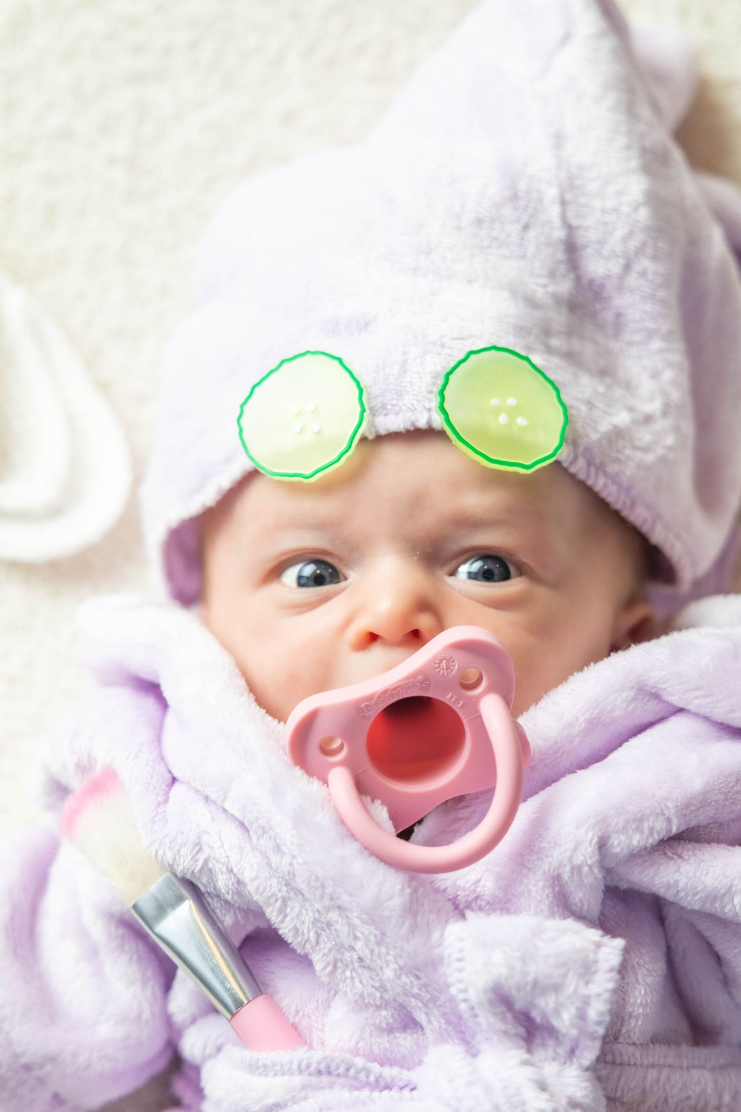 A baby wearing a spa-style towel wrap with cucumber slice decorations and a pink pacifier.