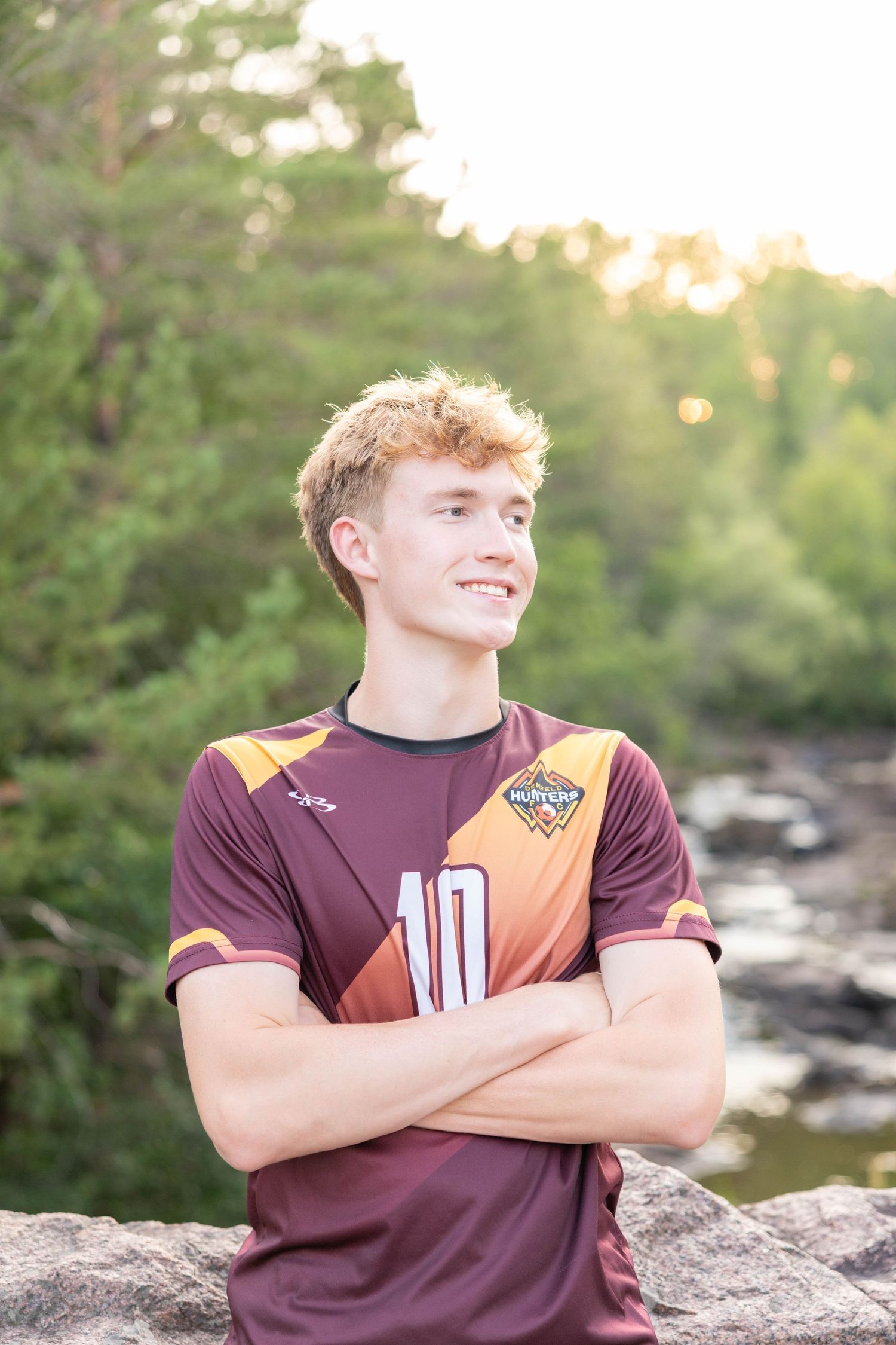 Teen boy in maroon soccer jersey number 10 stands arms crossed smiling outdoors near rocky river with trees.