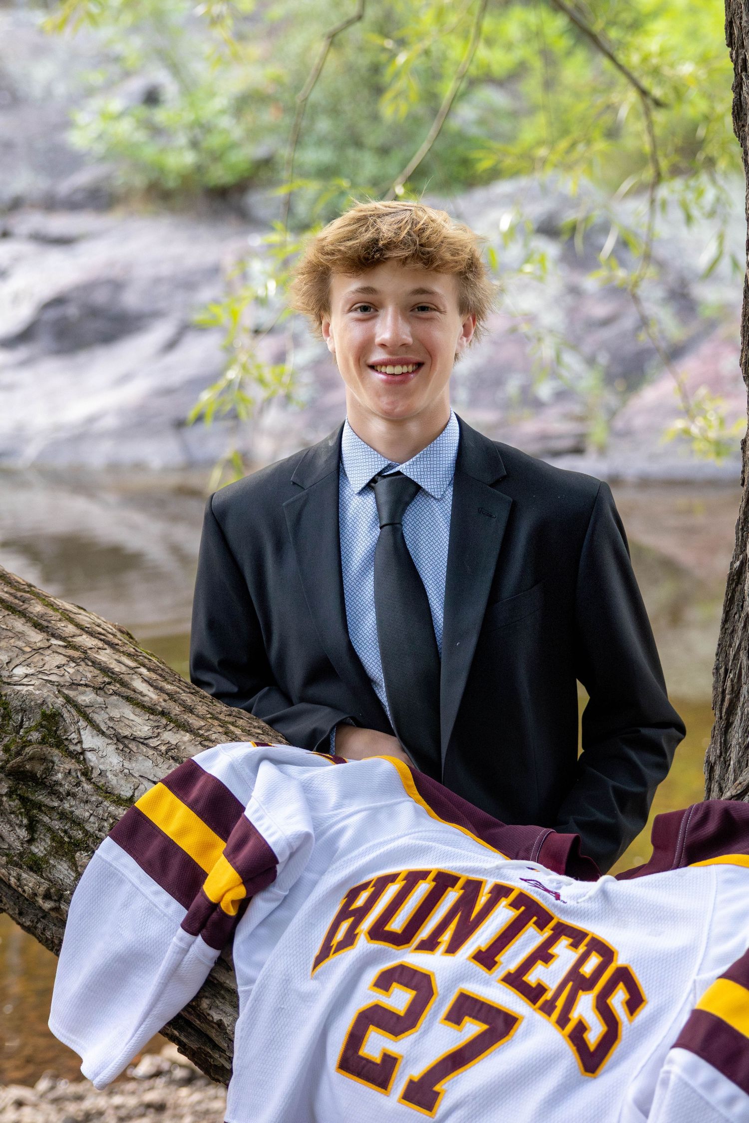 A young person in a black suit poses with a maroon and gold sports jersey against a natural outdoor background.