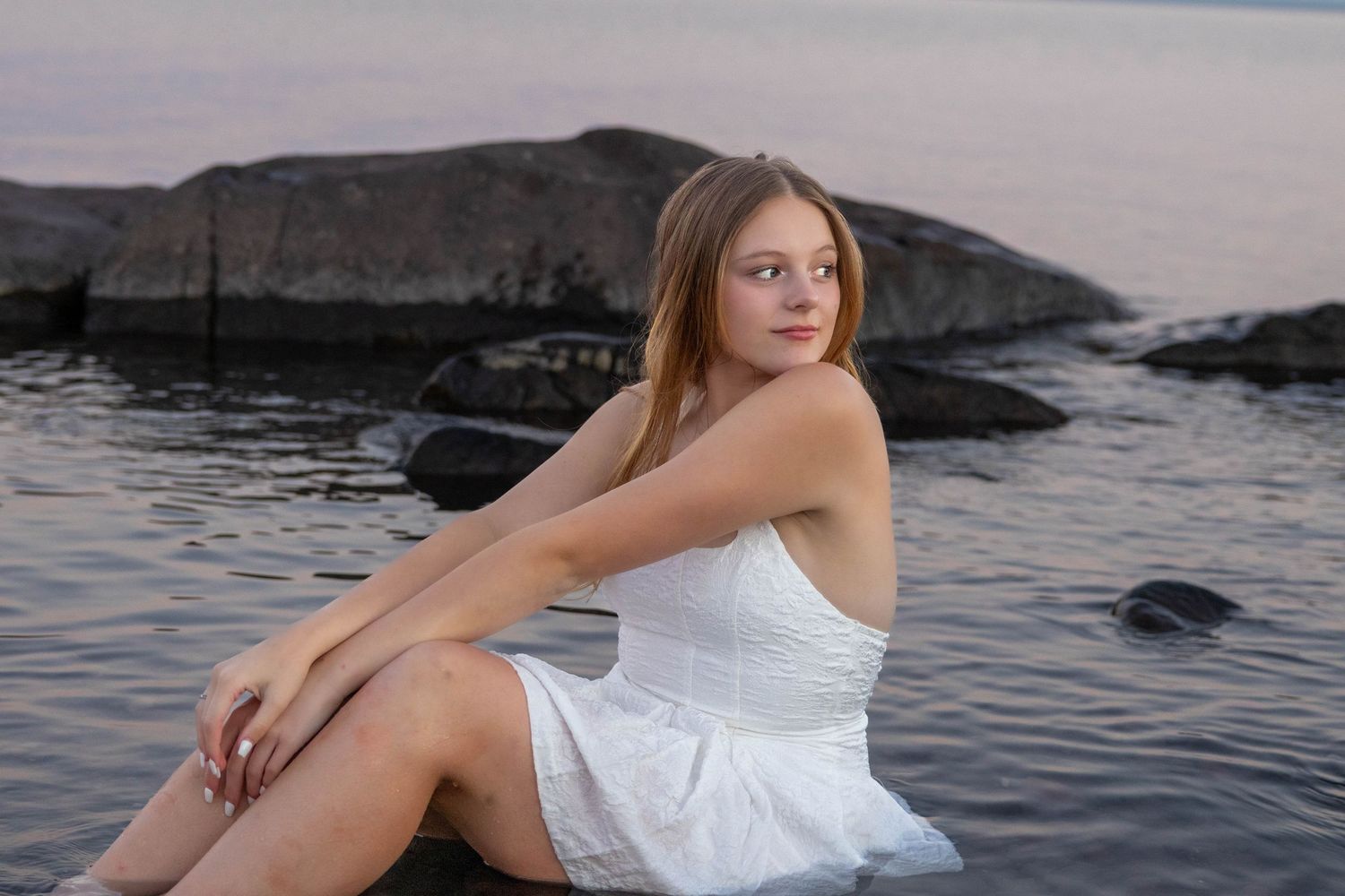 Person in white dress poses gracefully on rocky coastline during sunset photoshoot by the ocean.