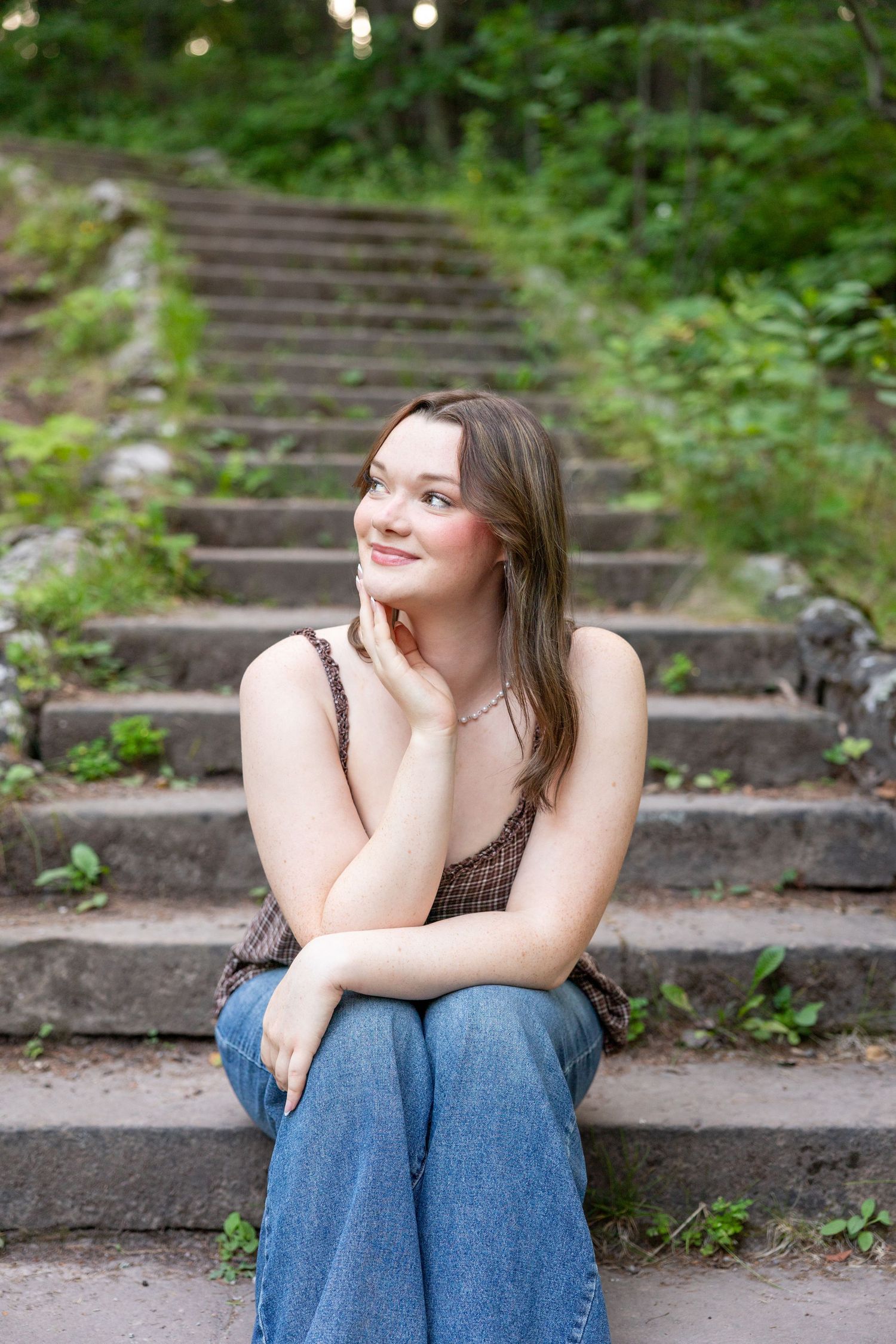 Young woman in brown top and jeans sits on stone steps, smiling thoughtfully with hand on chin, surrounded by greenery.
