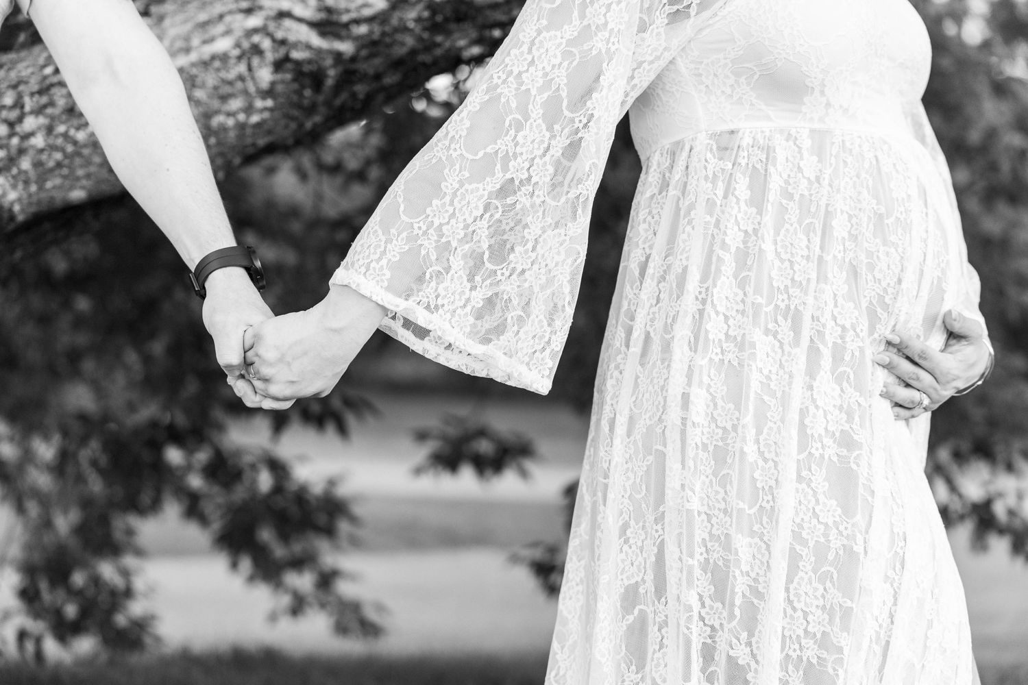 Black and white maternity photo of pregnant woman in lace dress holding hands with partner outdoors.